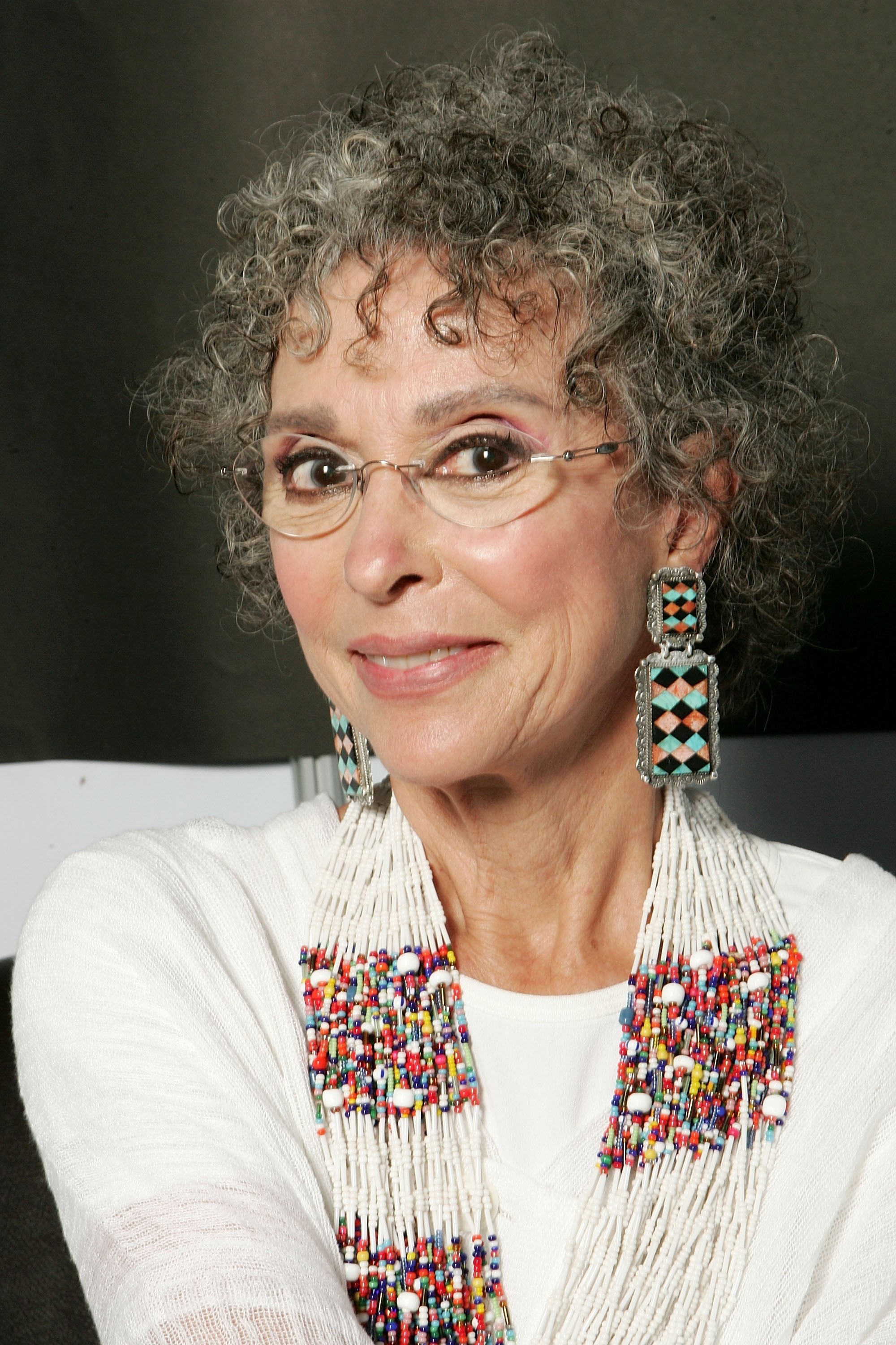An elegant woman with curly hair wears large beaded earrings and necklace, smiling softly at the camera
