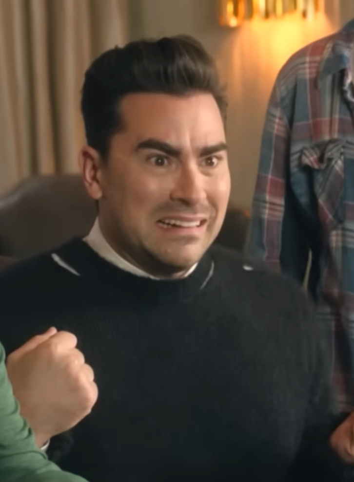 Daniel Levy with a surprised expression, wearing a dark sweater and collared shirt, with a clenched fist visible nearby, suggesting an emotional reaction