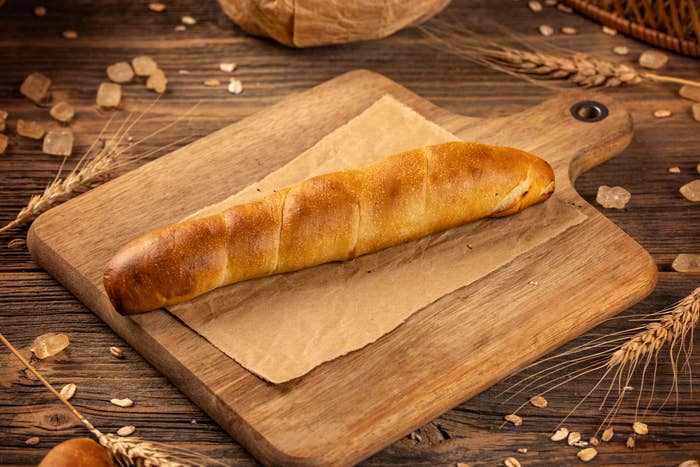 A freshly baked baguette on a wooden cutting board surrounded by scattered grains and wheat stalks