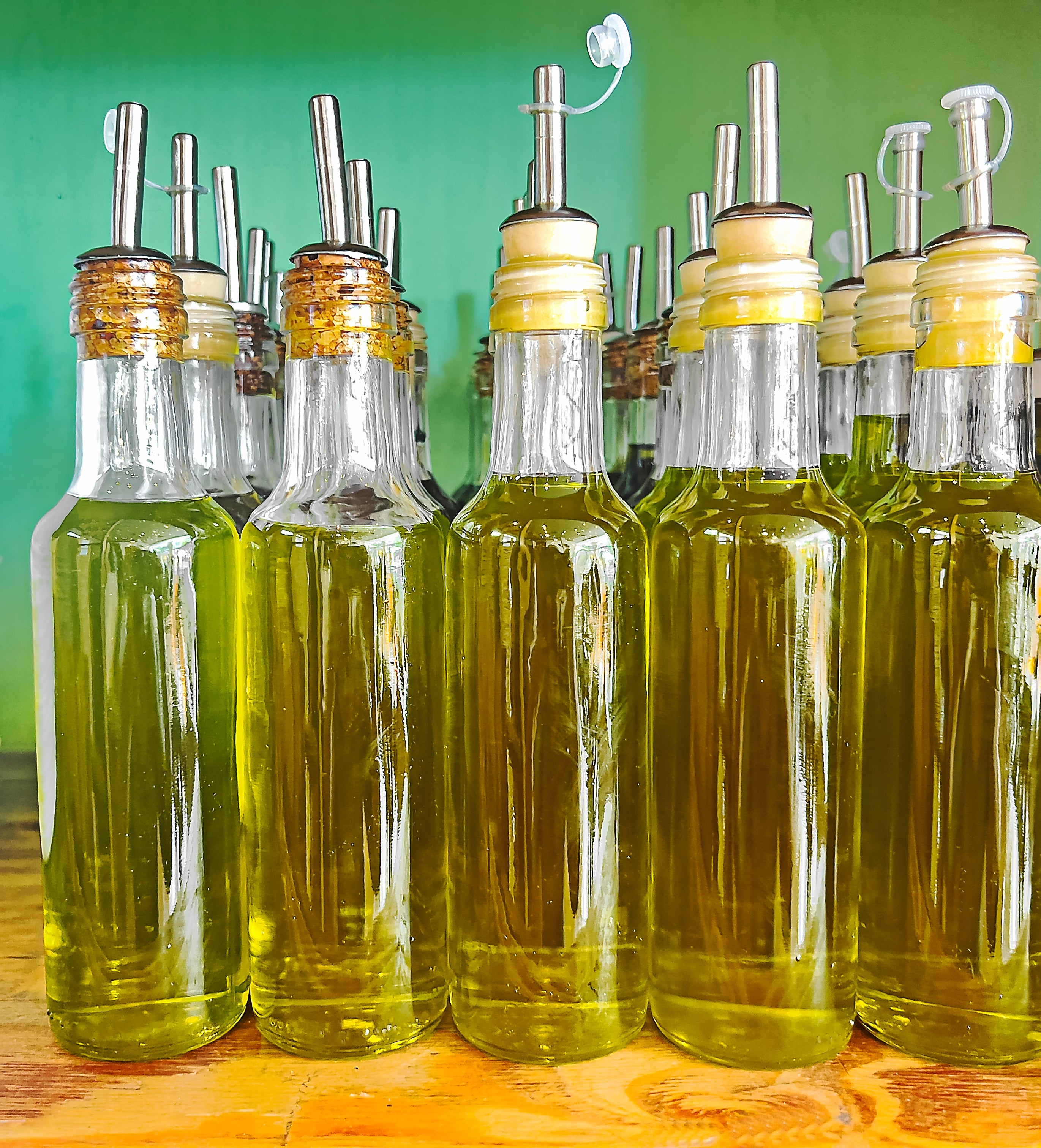 Rows of glass bottles filled with olive oil, each with a metal pour spout, placed on a wooden surface against a neutral background