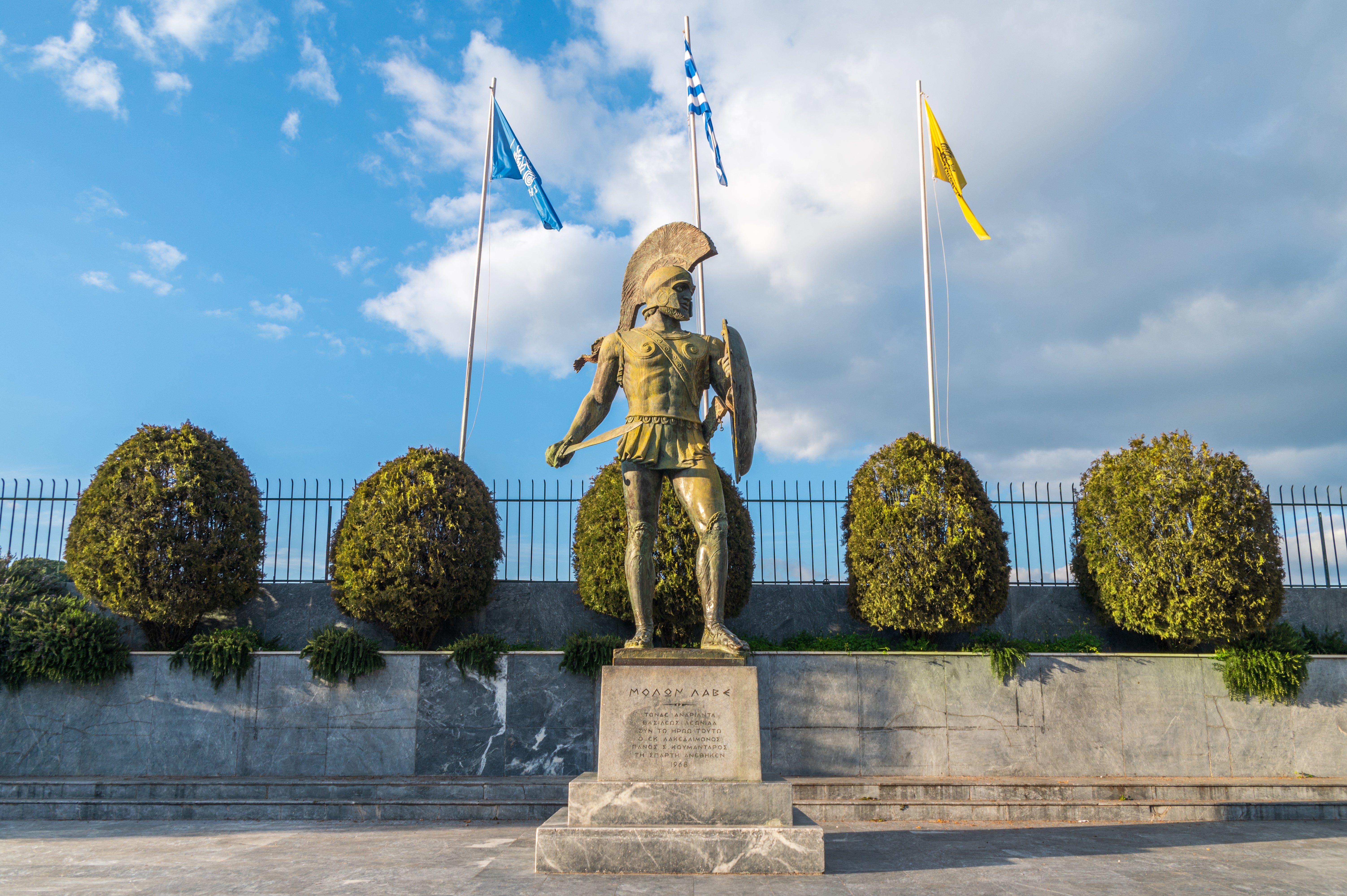 Statue of an ancient warrior with helmet and shield, surrounded by three flags on poles