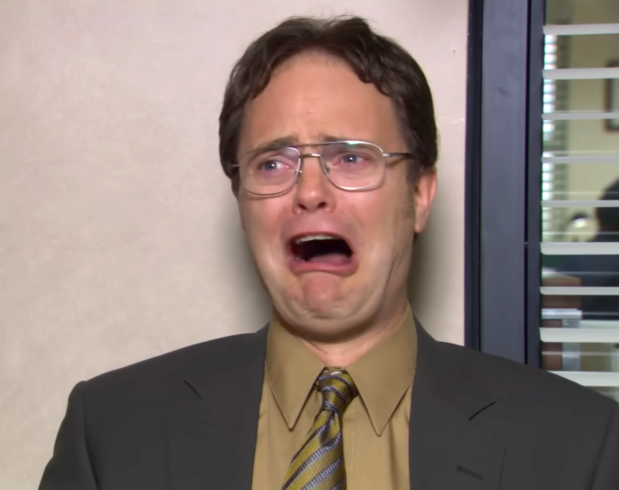 Rainn Wilson in an office setting with a distressed expression, wearing glasses, a suit, and a tie
