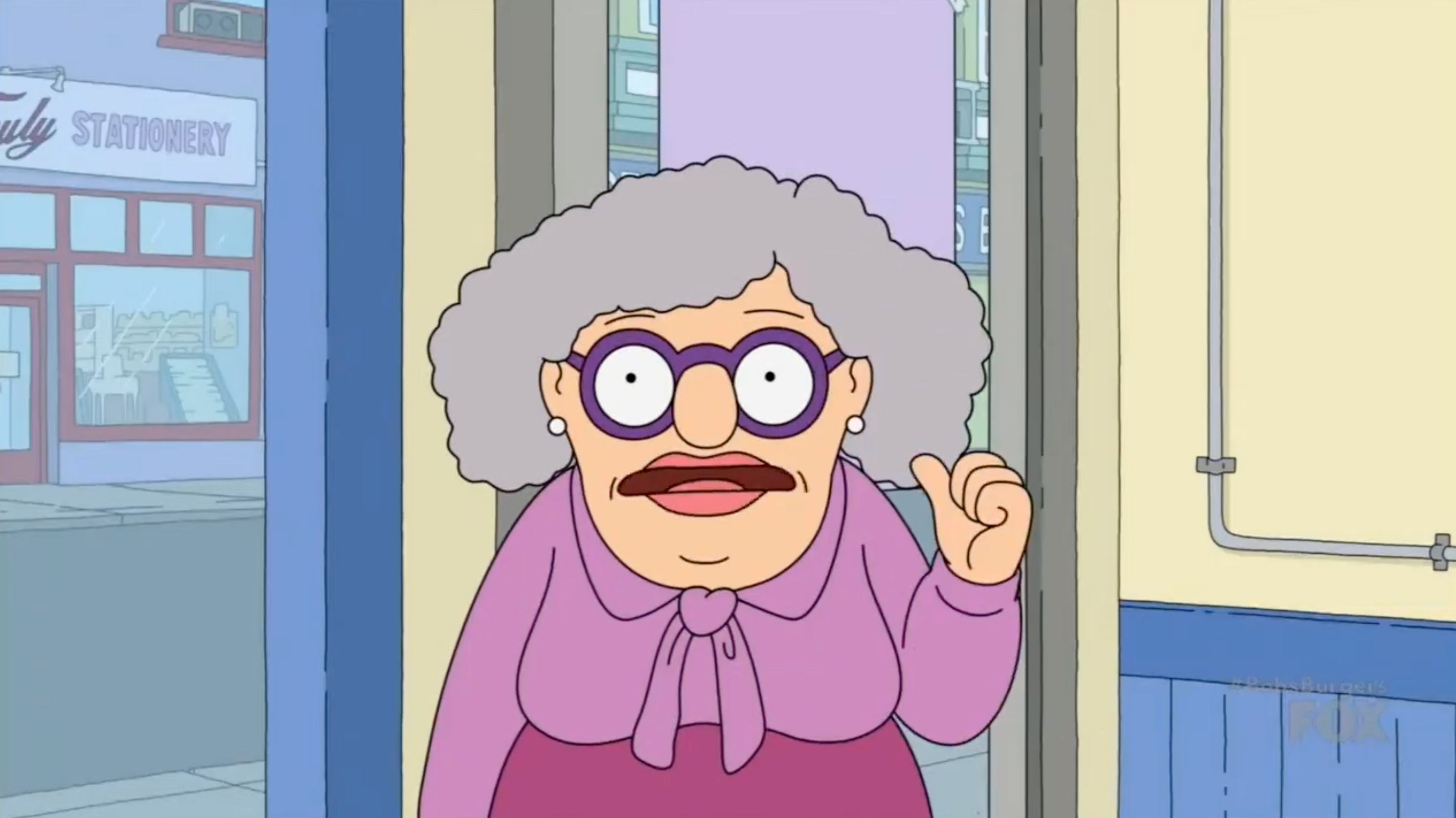 Animated older woman with curly hair, round glasses, and a purple bow-tie blouse gestures while standing indoors near a doorway