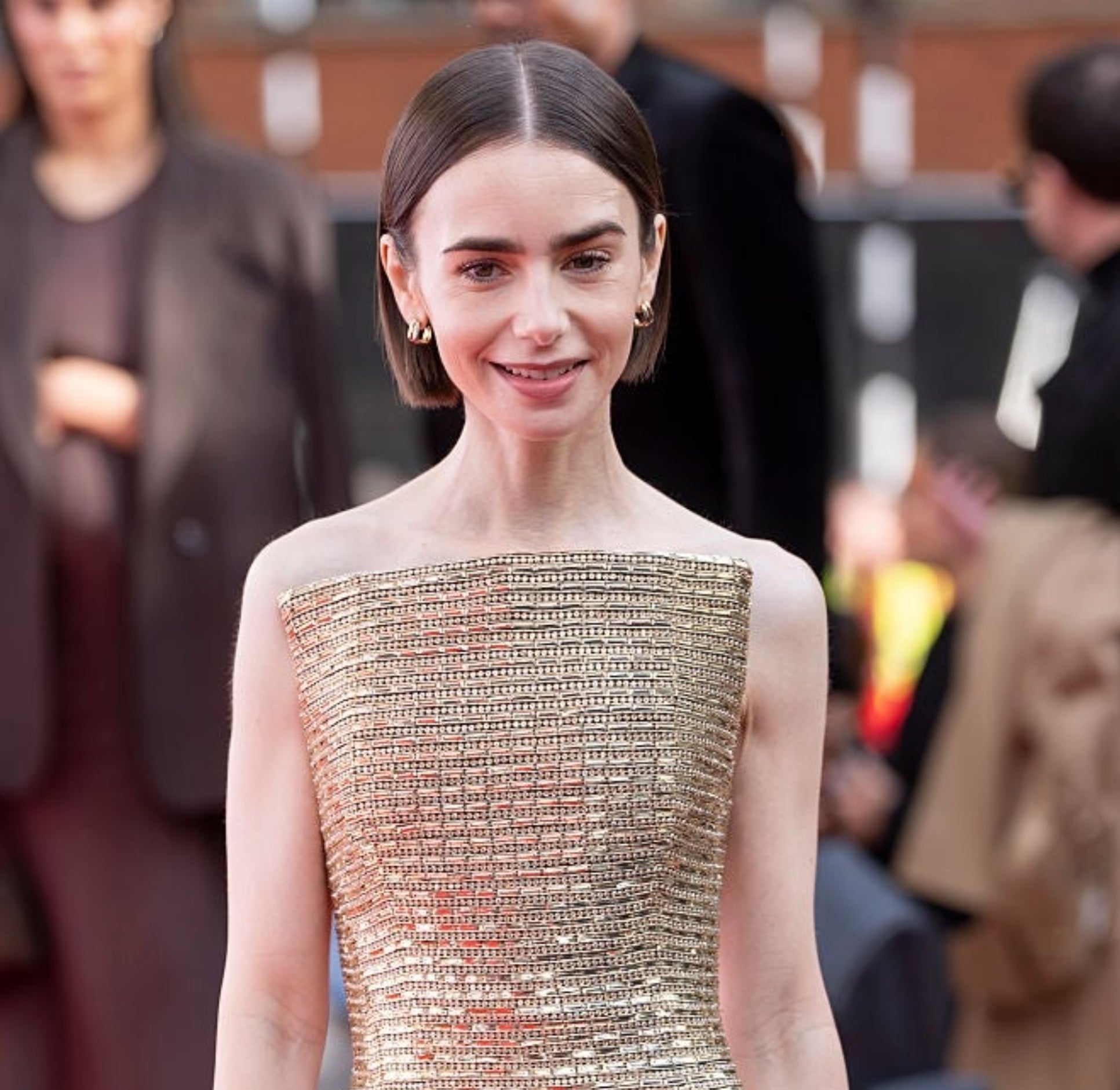 Person on red carpet in a form-fitting, textured gown with square neckline and short hair, smiling at the camera