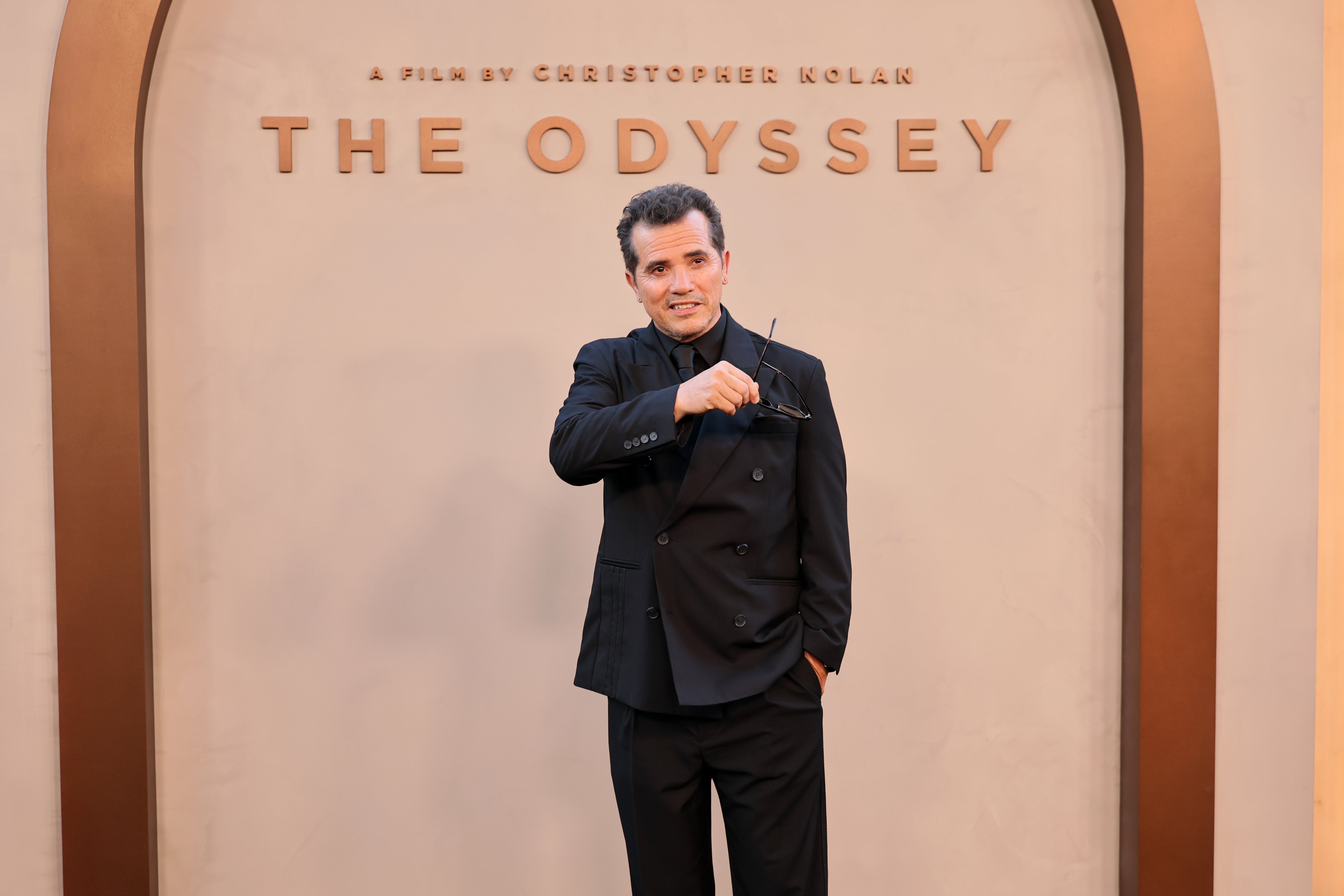 Person in a sleek black suit poses, holding glasses, at "The Odyssey" premiere with a backdrop arch and title text