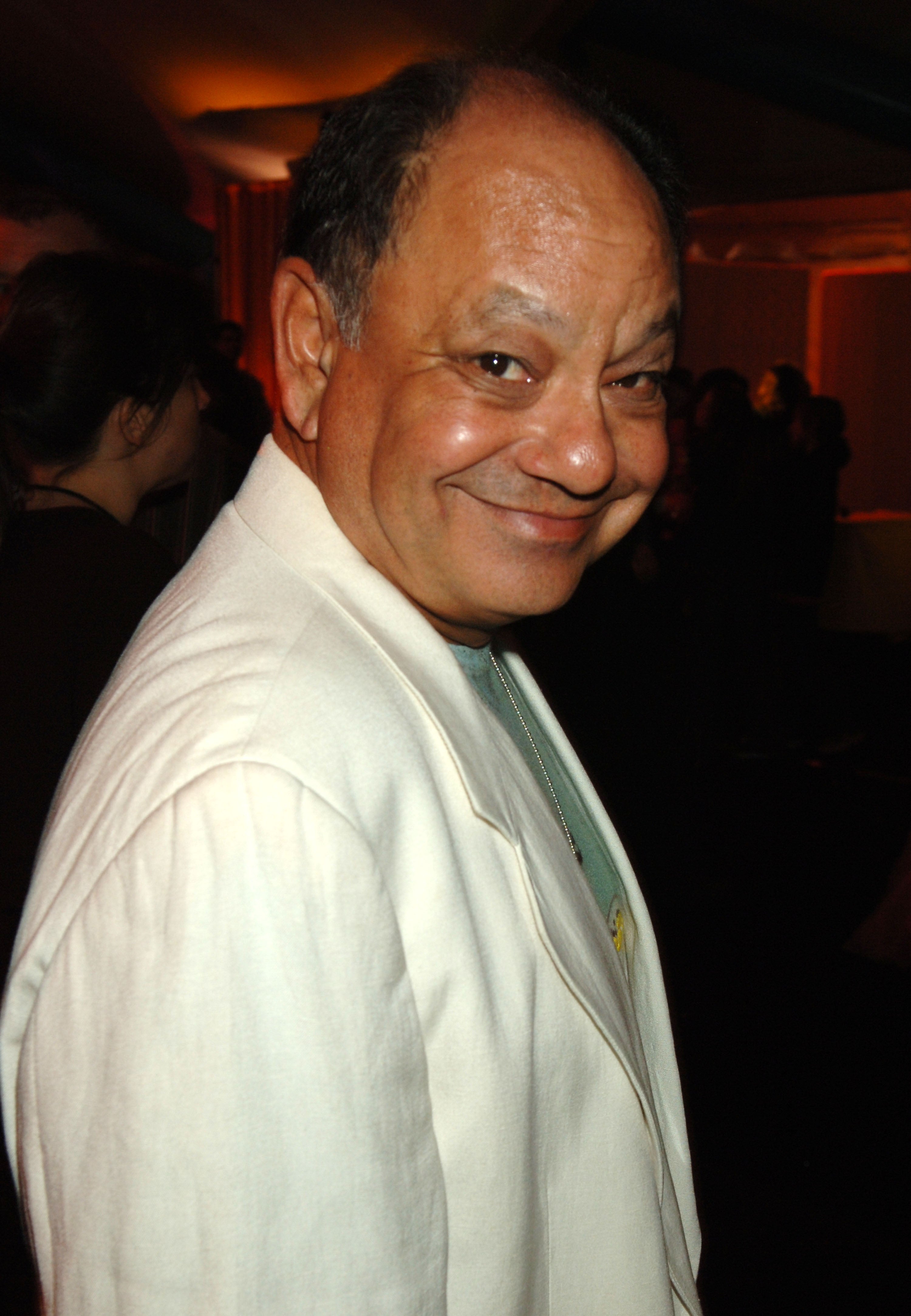 Man in a white blazer smiling slightly, standing in a dimly lit setting with blurred people in the background