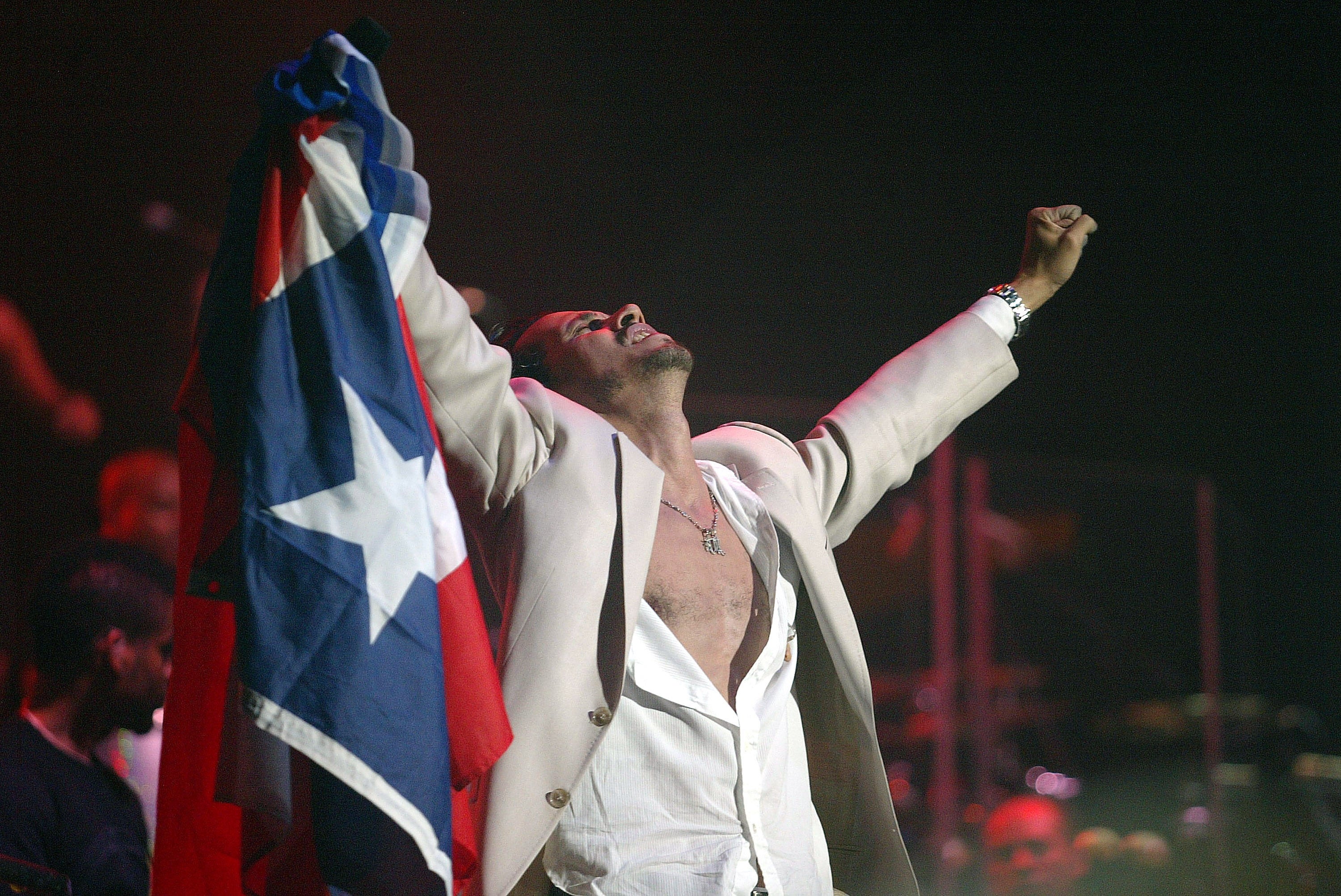 Person on stage holding a flag, wearing a partially unbuttoned shirt and jacket, celebrating energetically with raised arms