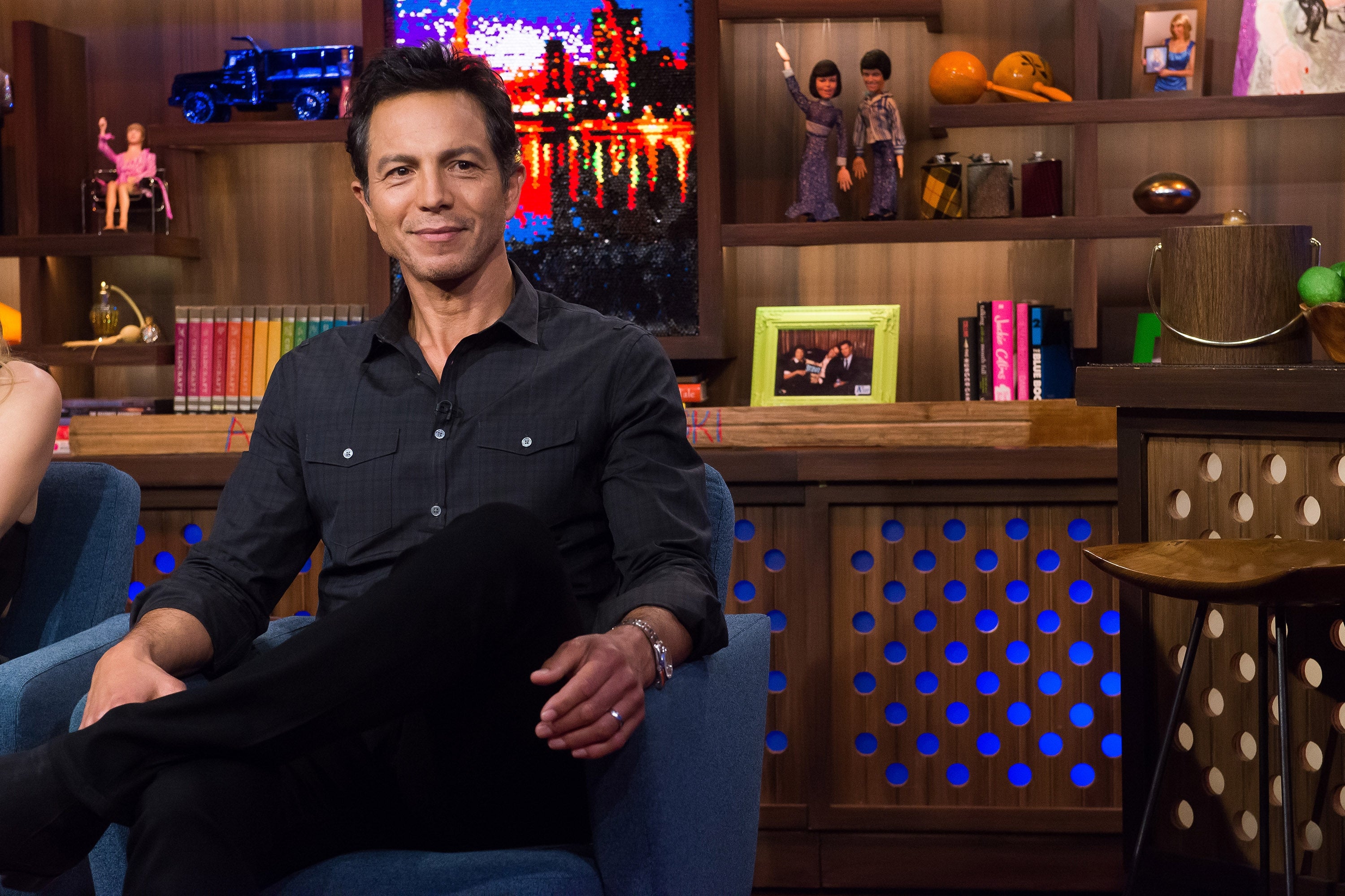 Person sitting in a TV studio, wearing a casual button-up shirt. Books and decor items are visible in the background