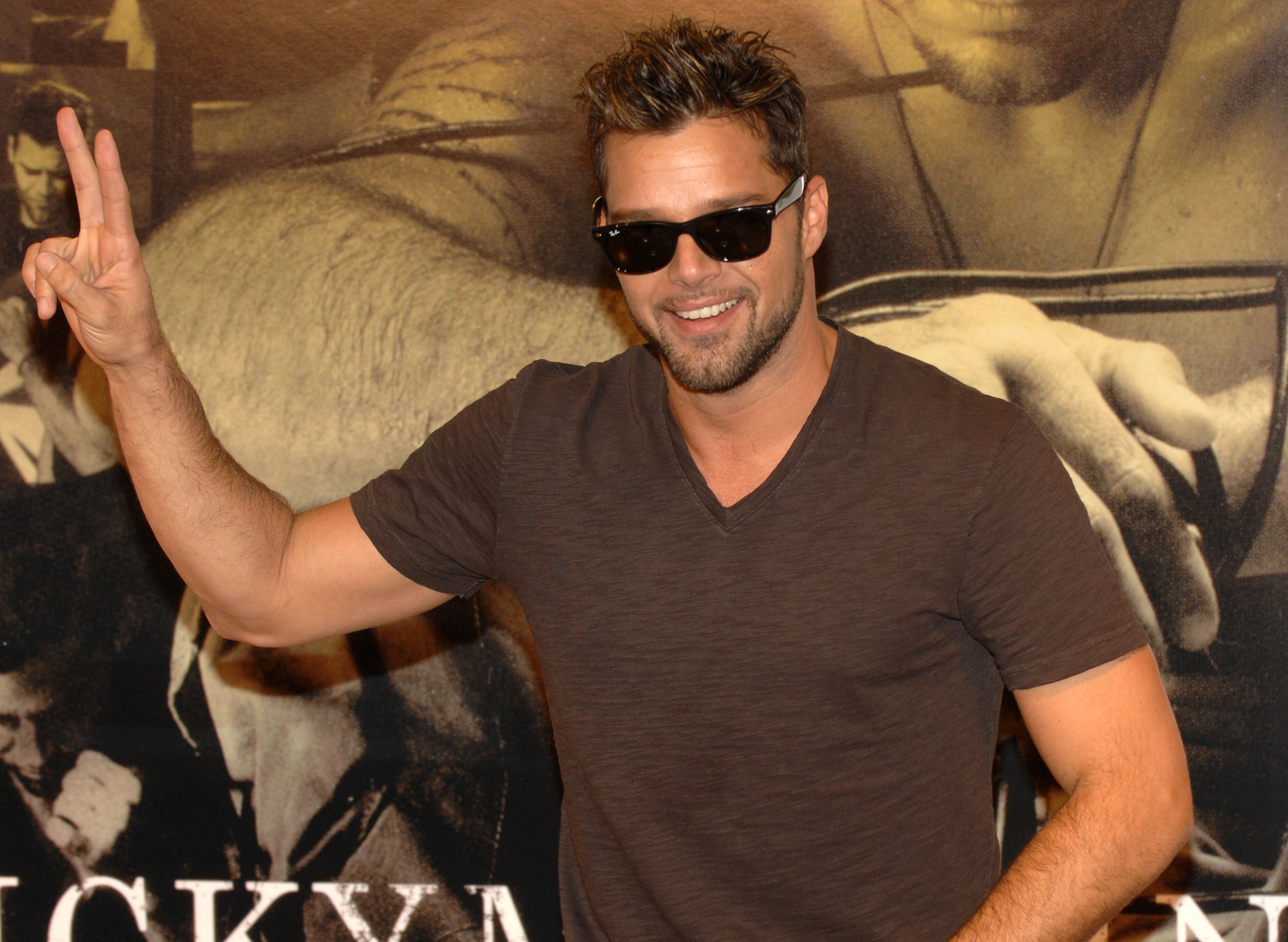 Man in sunglasses making a peace sign, wearing a casual T-shirt and smiling at an event with a poster background