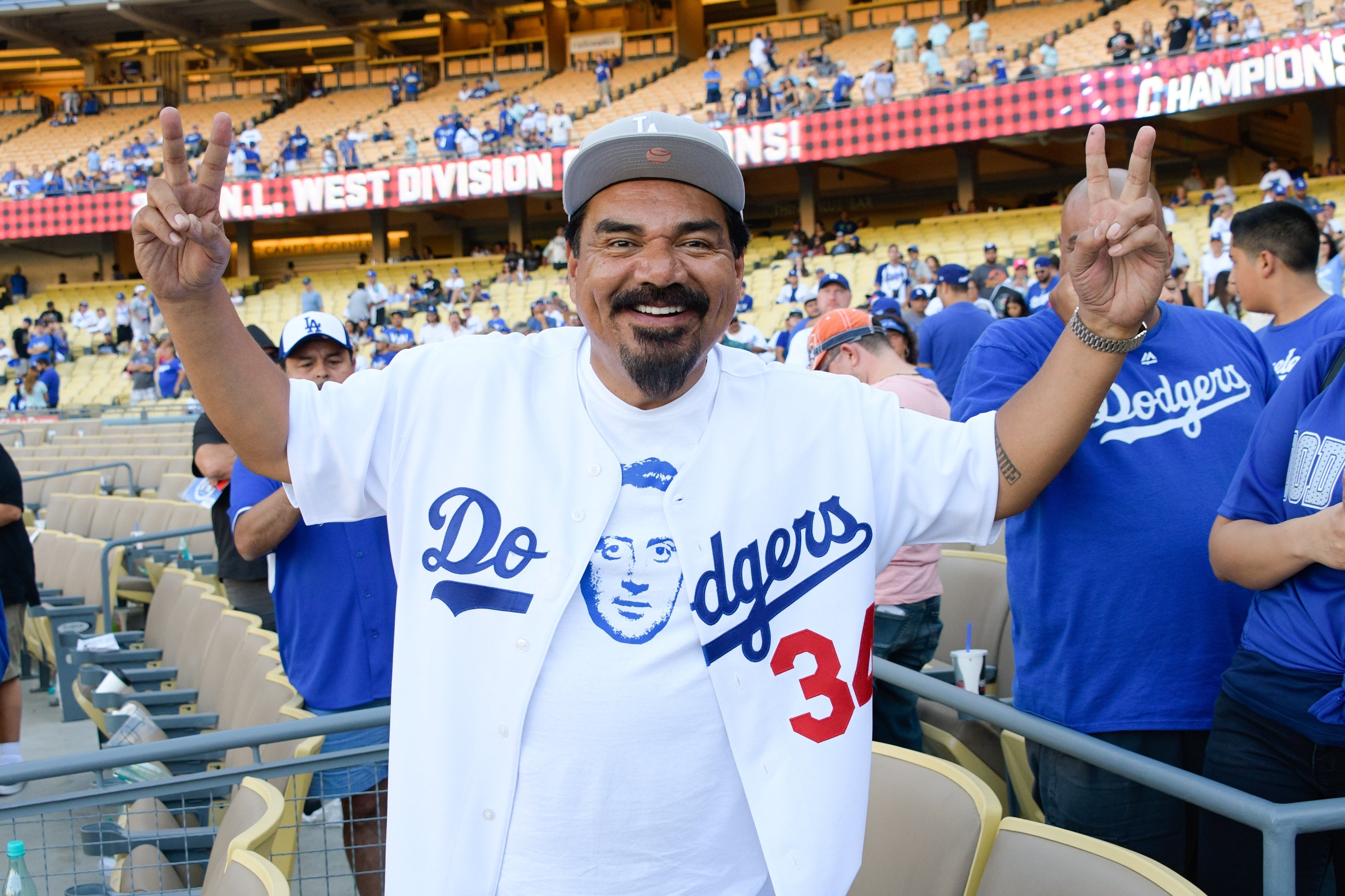 Person in Dodgers jersey and cap, smiling and holding up peace signs at a baseball stadium, surrounded by fans