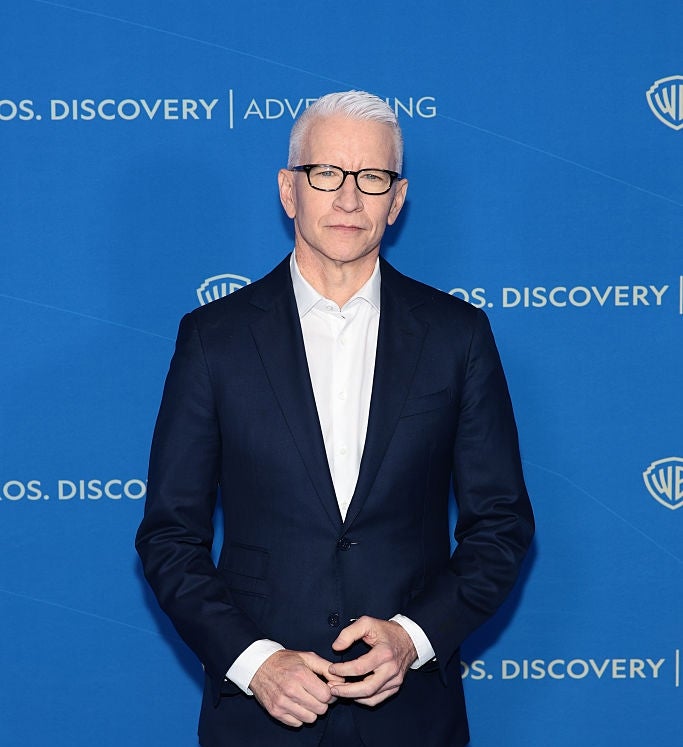 Person in a dark suit and white shirt standing against a branded backdrop at a media event