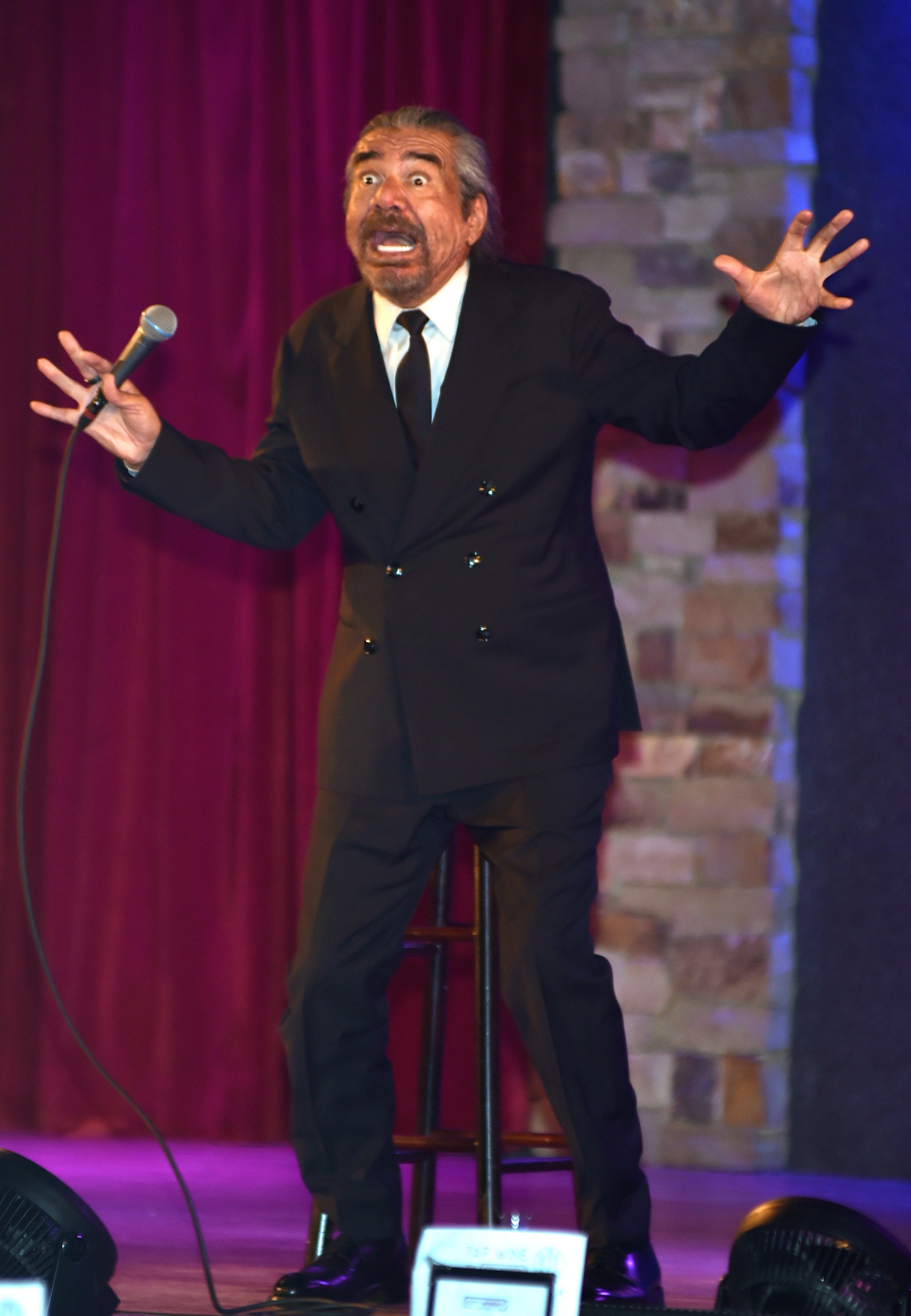 A man in a double-breasted suit performs stand-up comedy on stage, holding a microphone with an animated expression and gestures