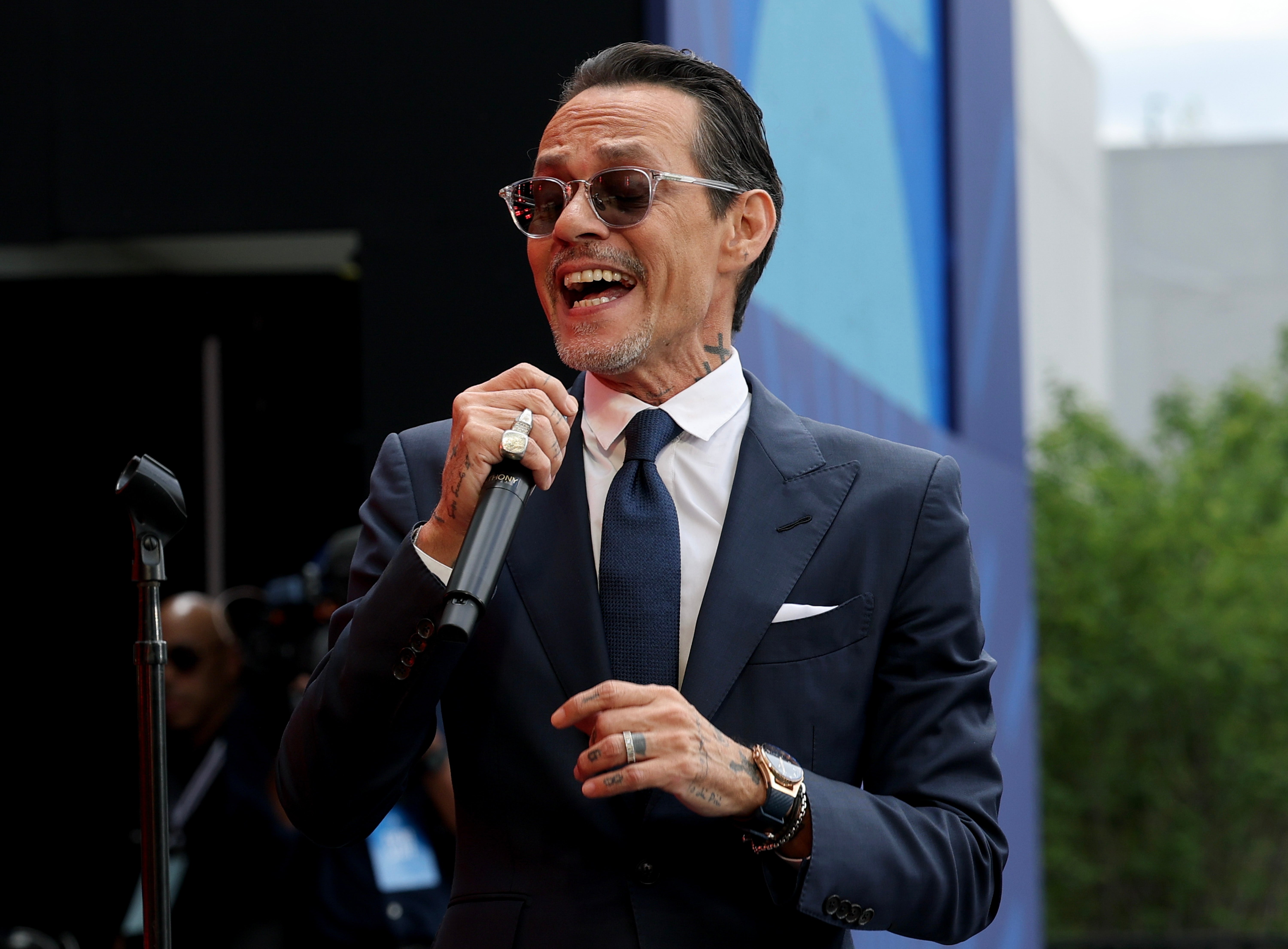 A man in a suit and tie performs passionately on stage holding a microphone, wearing sunglasses and rings