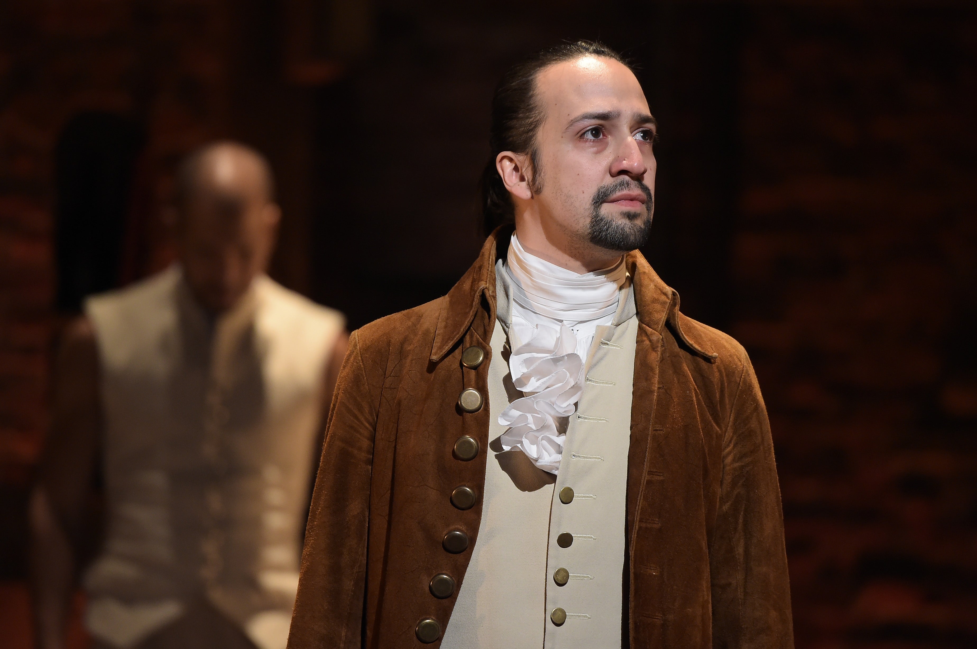 A person in period costume with a ruffled shirt and coat stands on stage, appearing thoughtful. Another person is blurred in the background