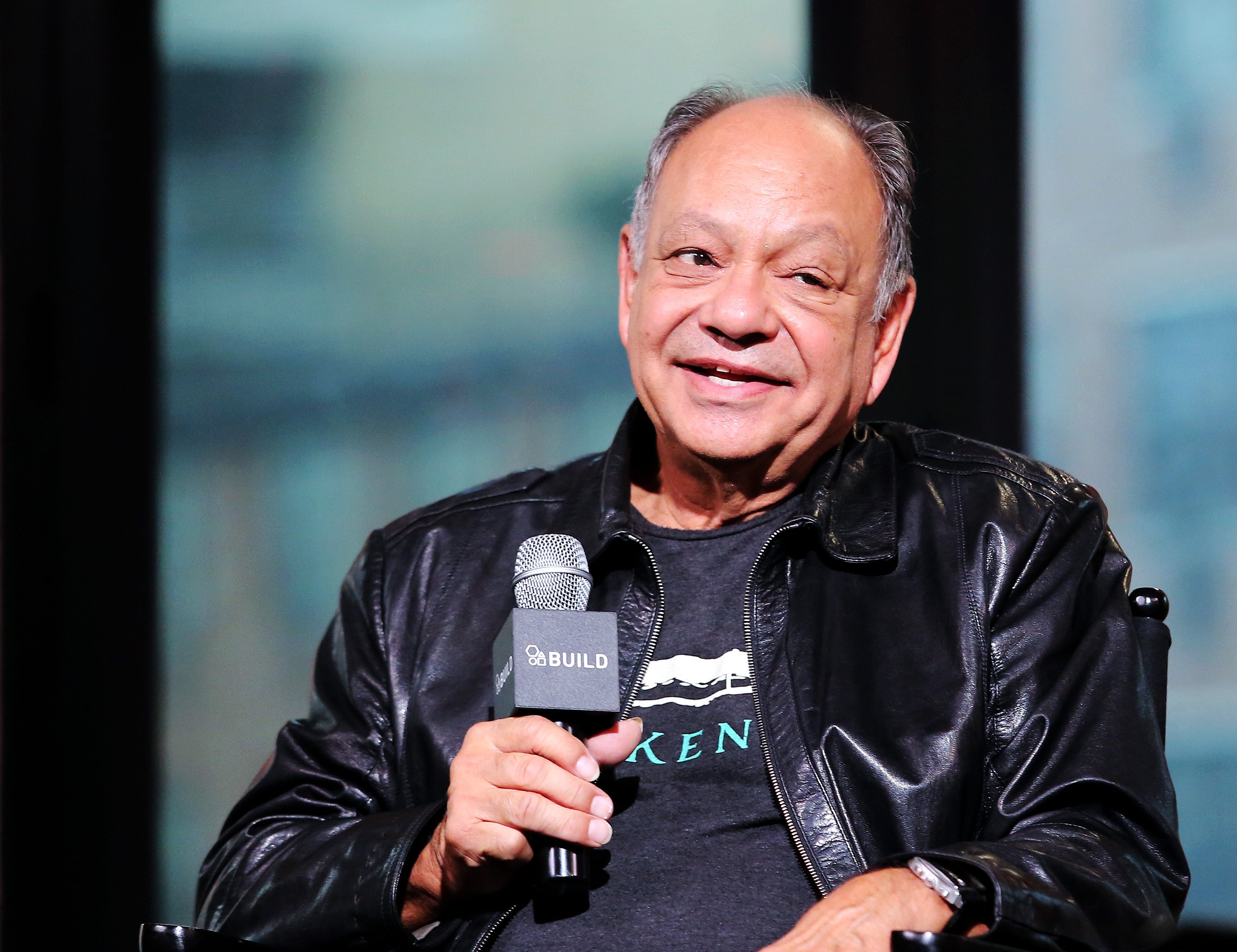 An individual in a leather jacket holds a microphone with a "BUILD" logo, seated and speaking at an event