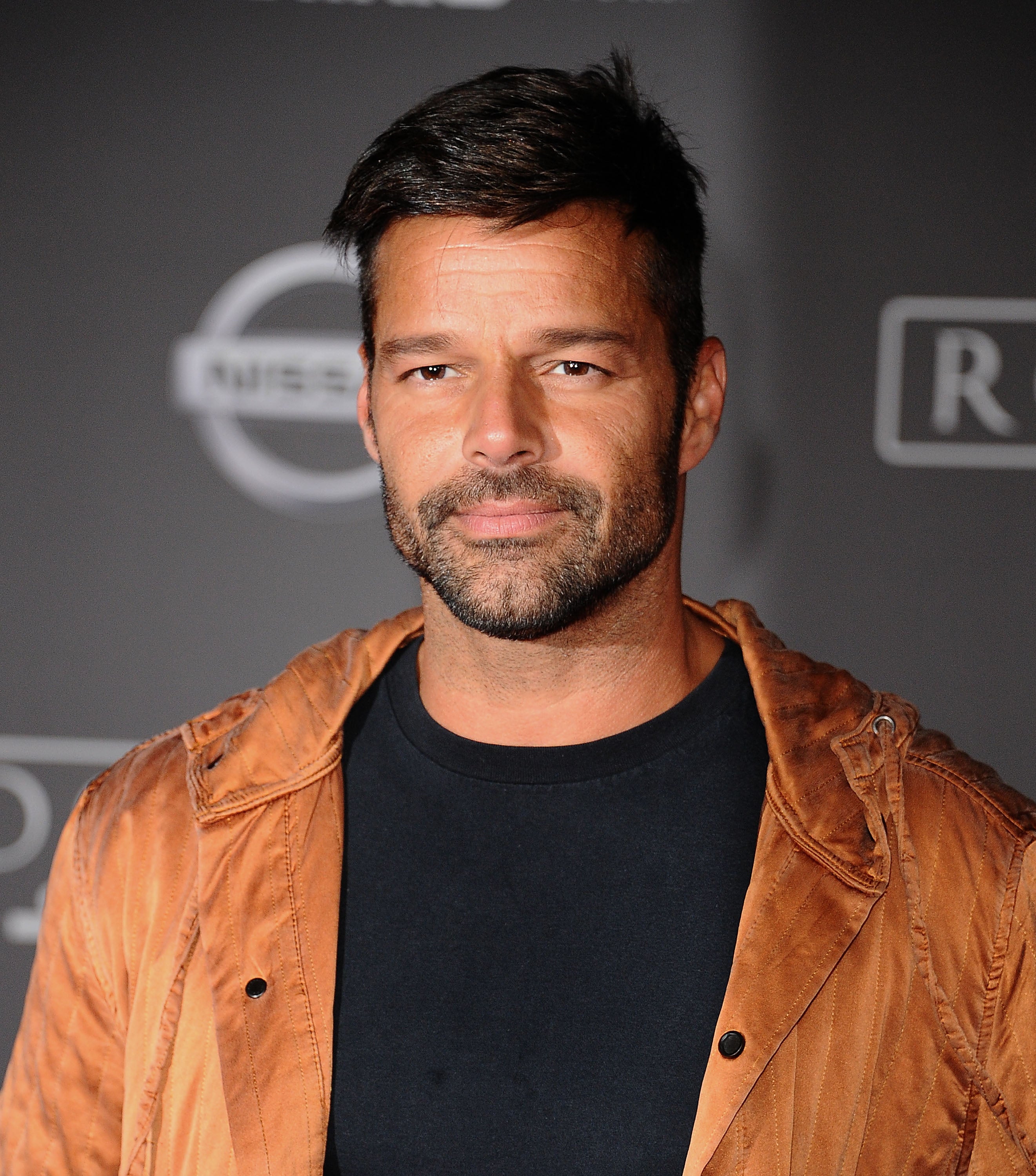 A person poses at an event wearing a casual black shirt under a stylish brown jacket, with a neutral backdrop