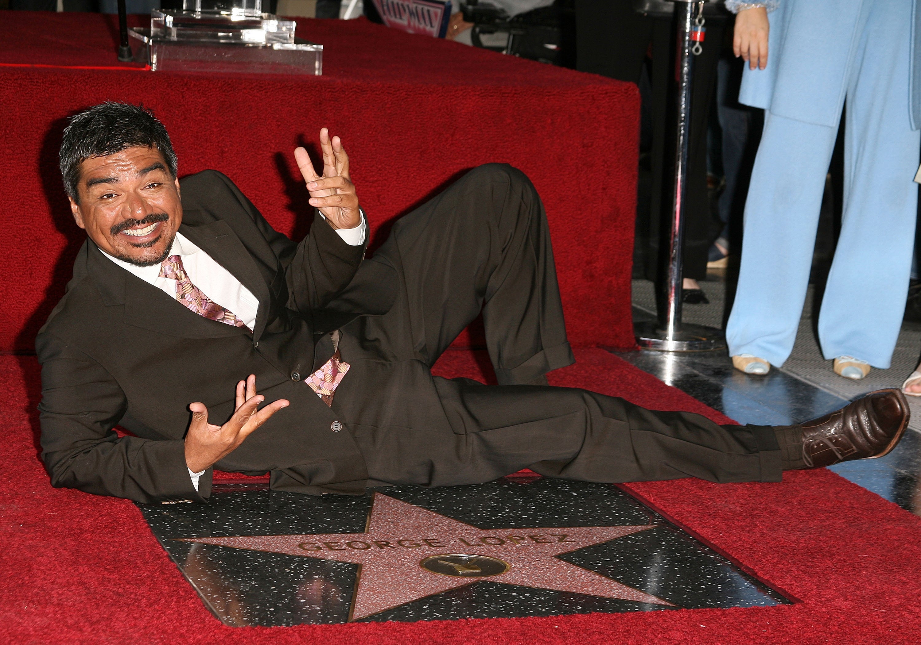 Person playfully poses on a Hollywood Walk of Fame star with their name engraved. Wearing a suit with a patterned tie