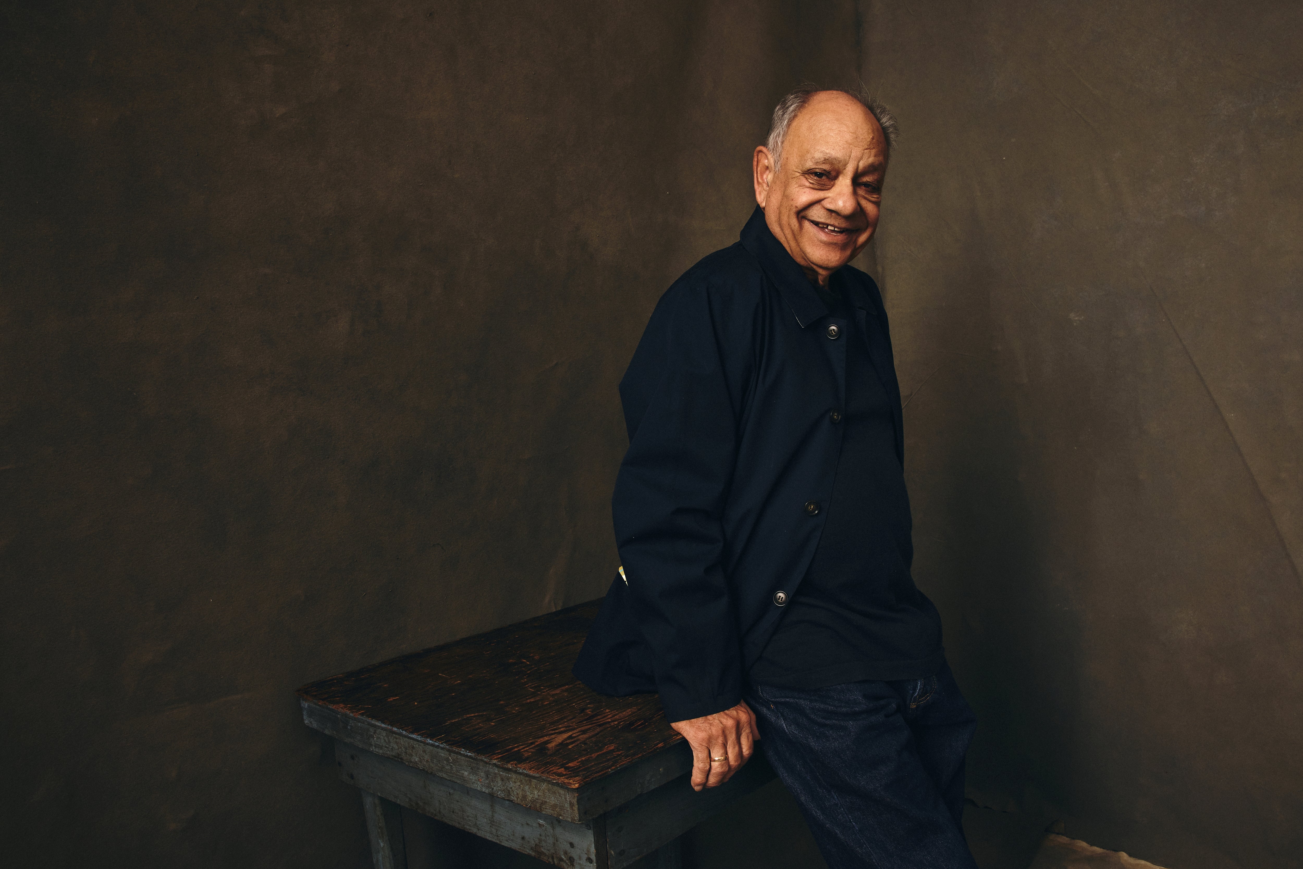 An older man sits on a wooden table, wearing a dark jacket and jeans, smiling warmly at the camera against a simple backdrop