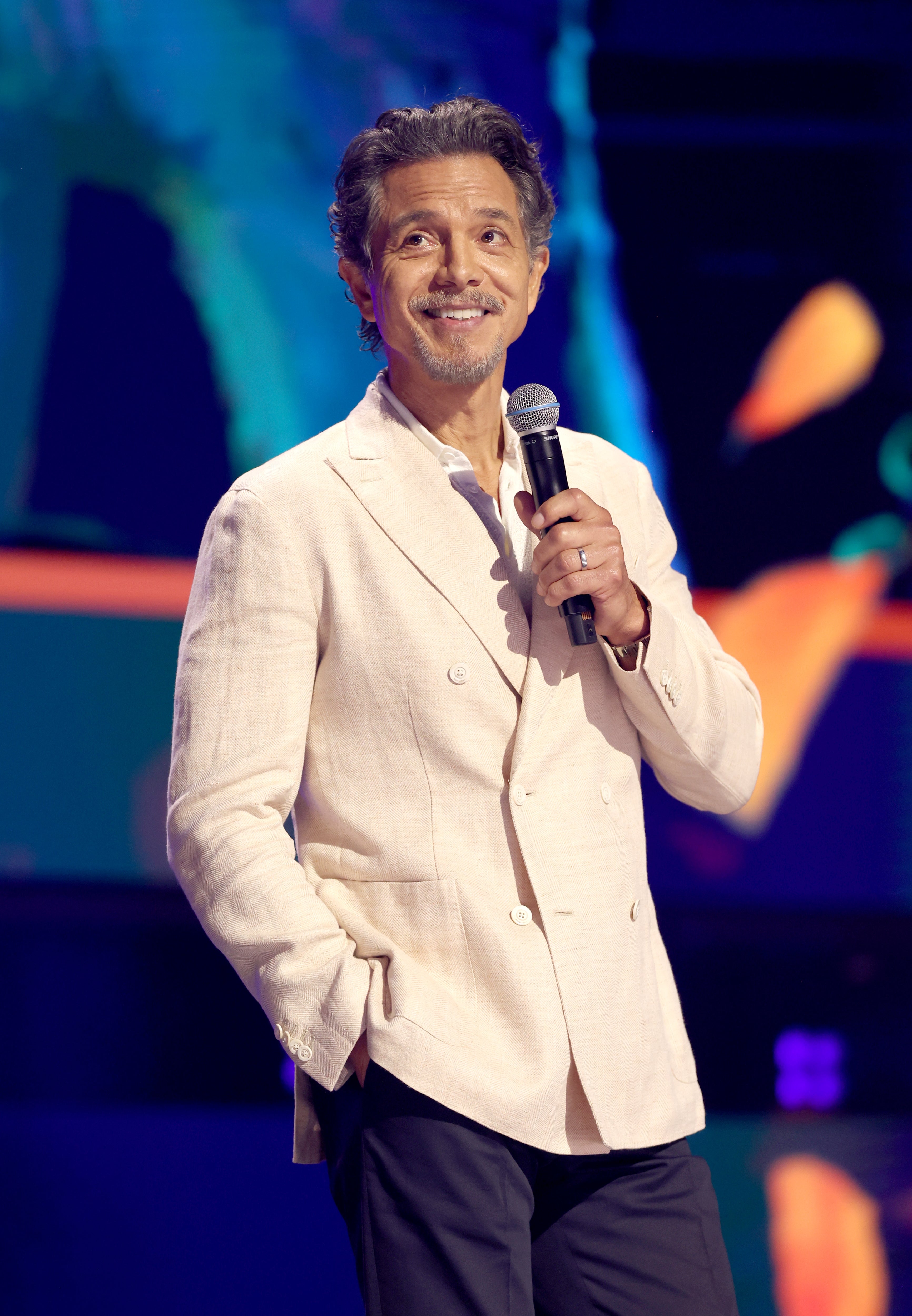 Person smiling on stage, holding a microphone, wearing a light blazer and dark pants, against a colorful background