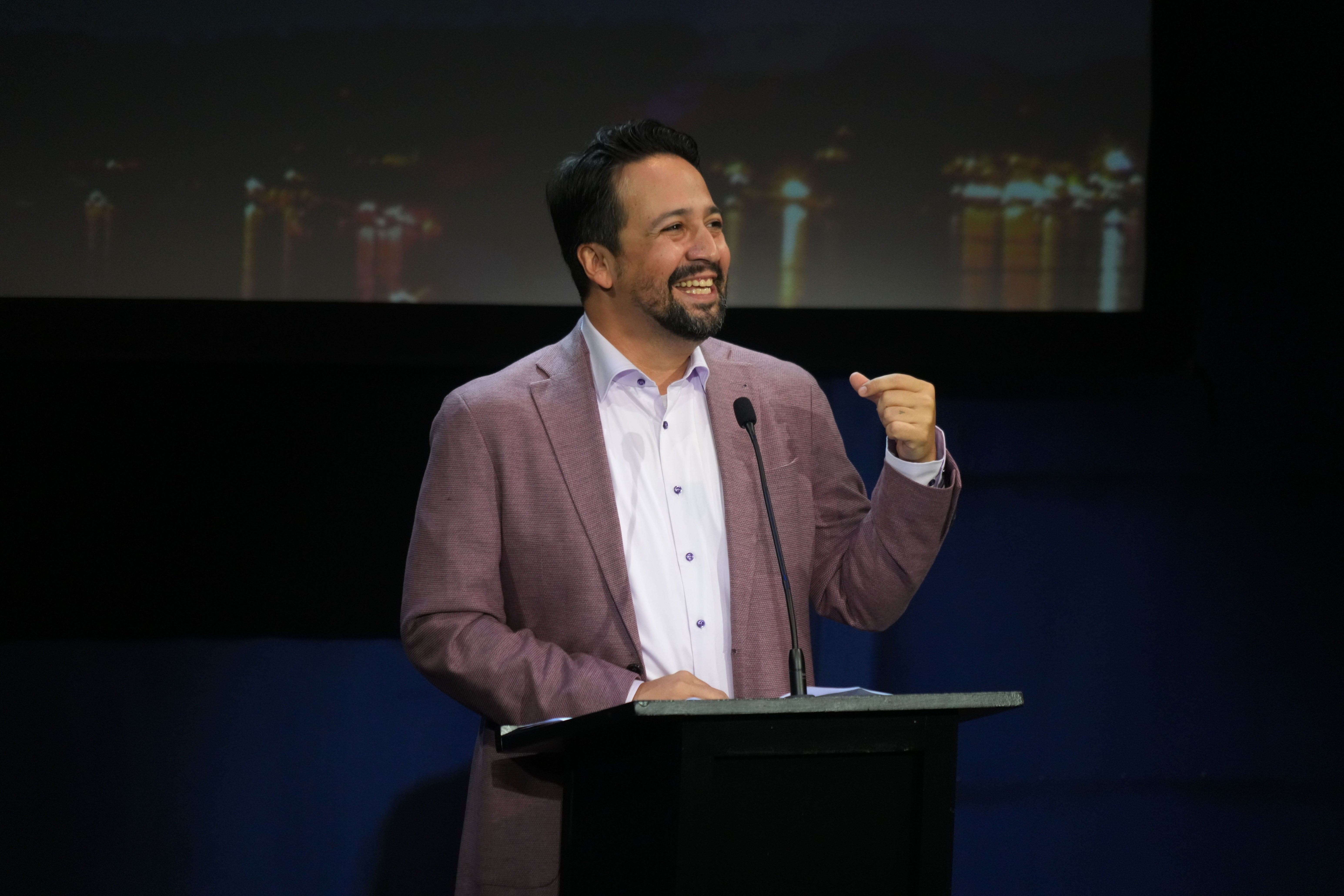 A person in a blazer speaks at a podium, smiling