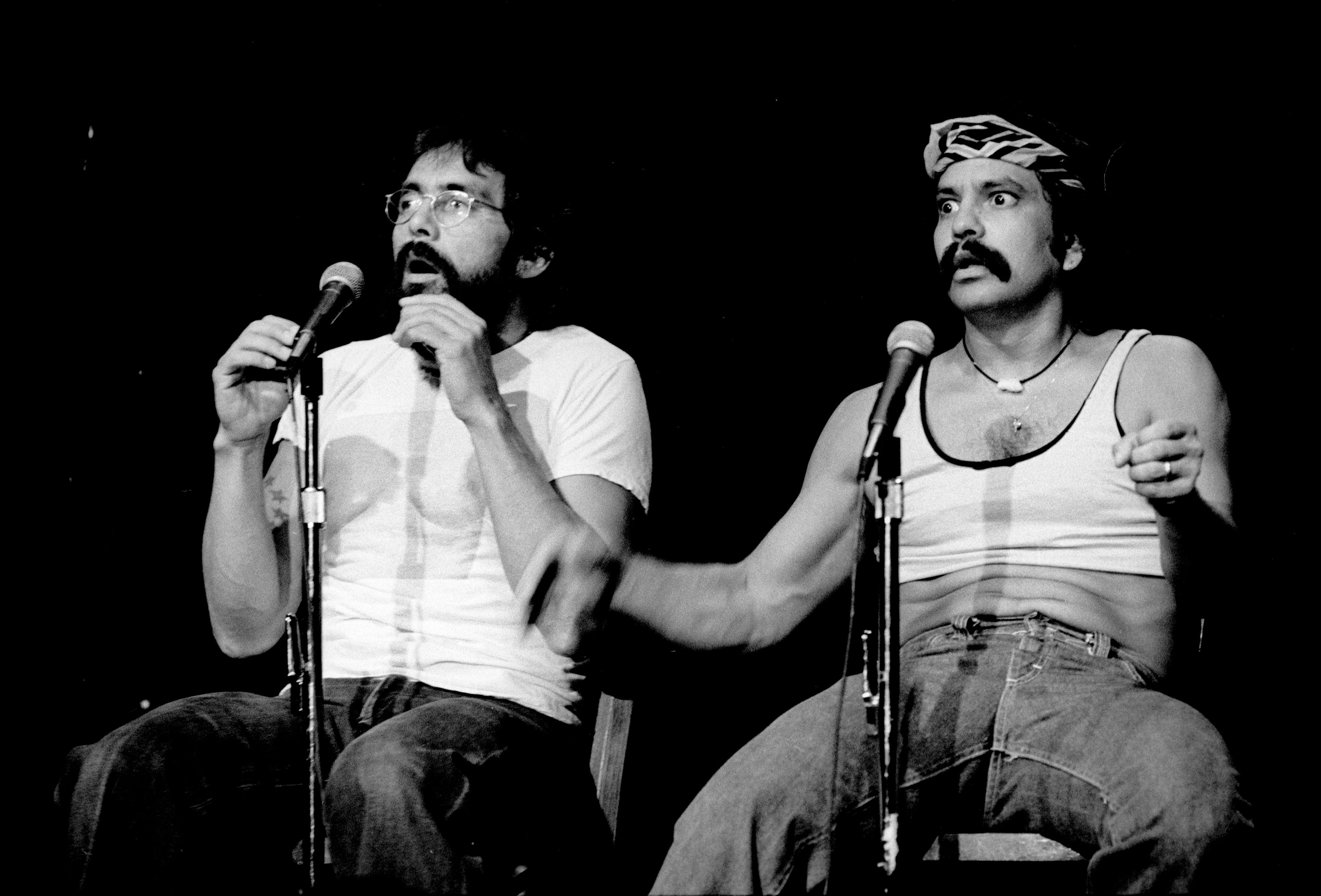Two performers sit on stage, each holding a microphone. One wears a bandana and tank top, the other wears glasses and a T-shirt