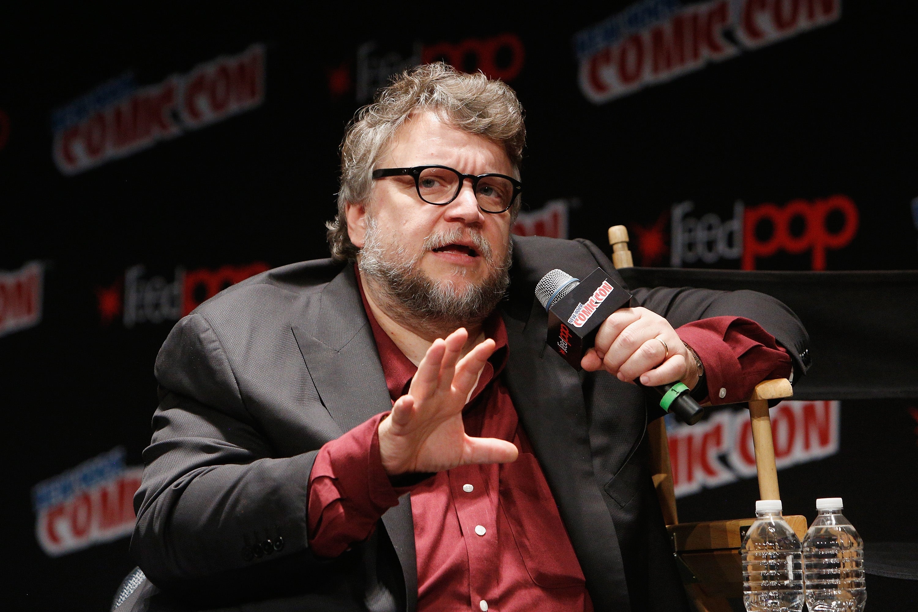 A person speaks into a mic at a Comic Con panel, wearing a suit jacket over a casual shirt, gesturing with one hand