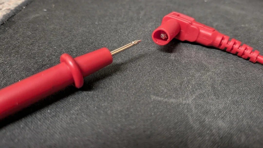 Close-up of pointed red multimeter probe next to its red connector on a dark surface