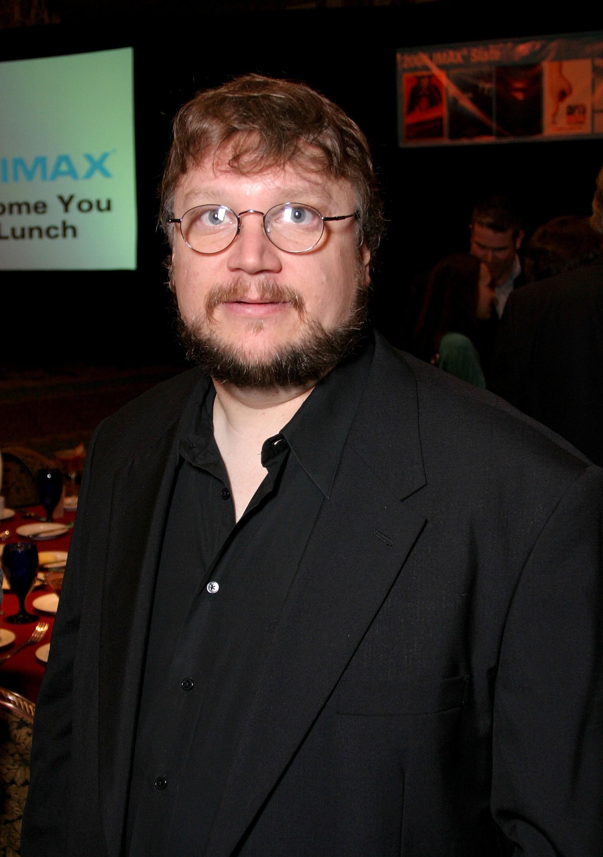 Person in black suit at an indoor event, standing near tables with people in the background