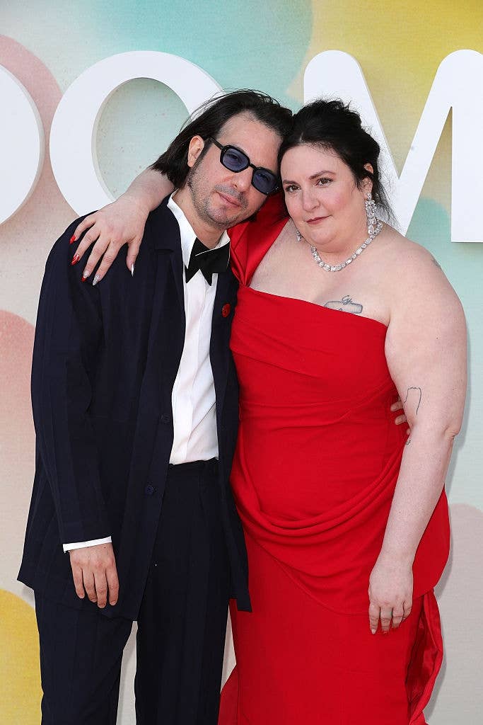 Two individuals pose together; one in a suit with sunglasses and the other in a strapless gown with a necklace