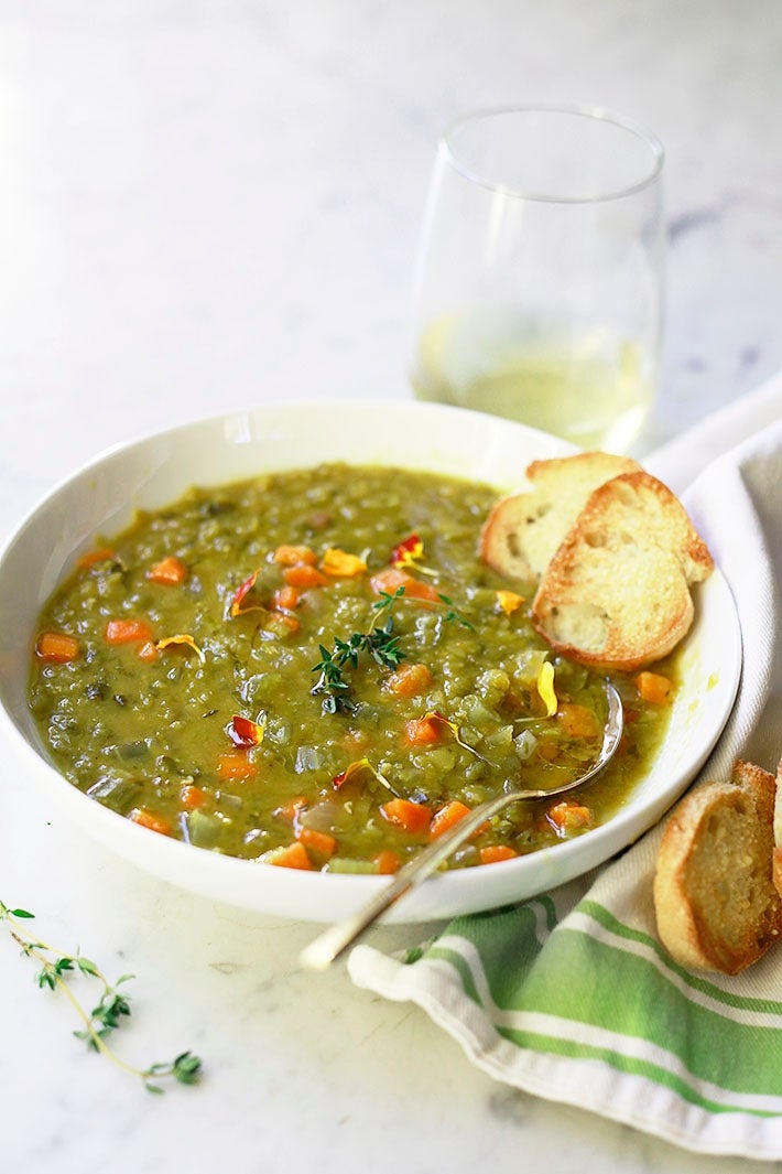 A bowl of split pea soup with diced vegetables, garnished with herbs, next to toasted bread and a glass of white wine on a table