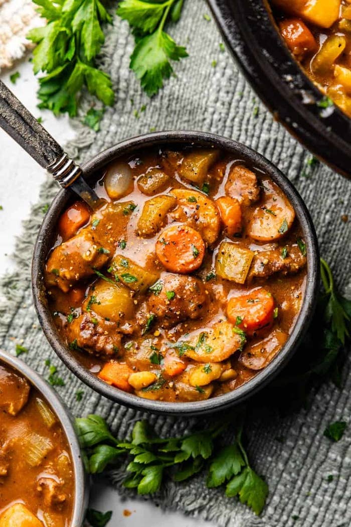 Hearty stew in a bowl with chunks of meat, potatoes, carrots, and herbs, sitting on a textured fabric next to fresh parsley