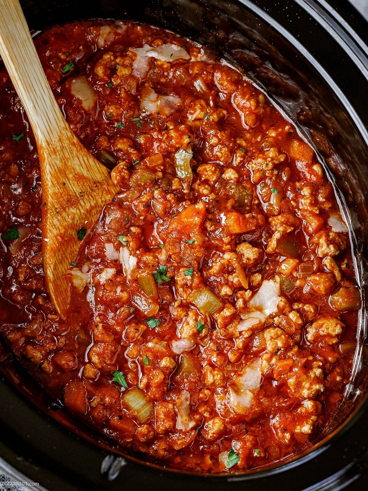 Hearty chili with ground meat, chopped vegetables, and tomato sauce in a slow cooker with a wooden spoon