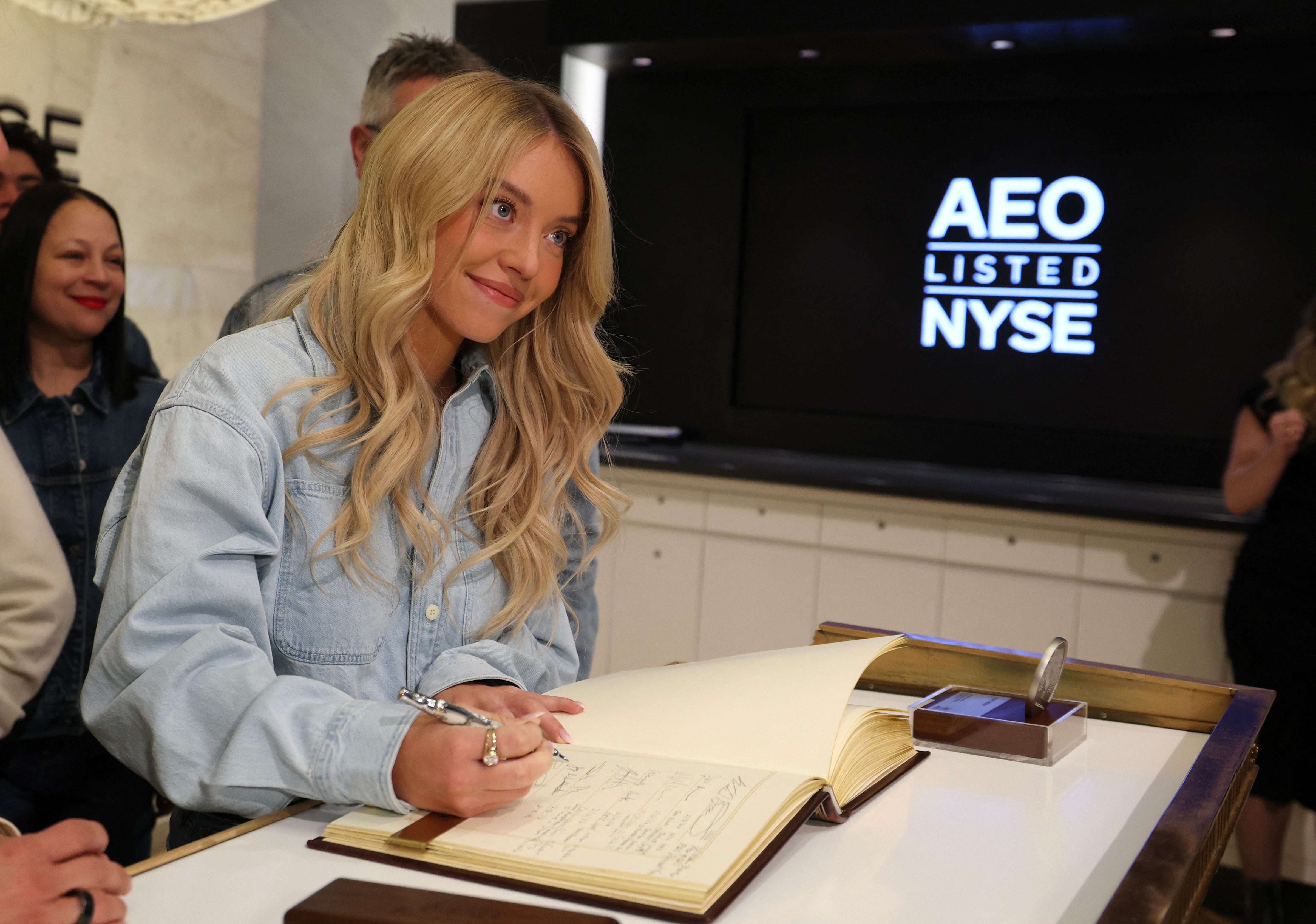 Person som signerer en bok på et NYSE-arrangement. De har på seg et uformelt denimantrekk, med mennesker og et AEO NYSE-skilt i bakgrunnen