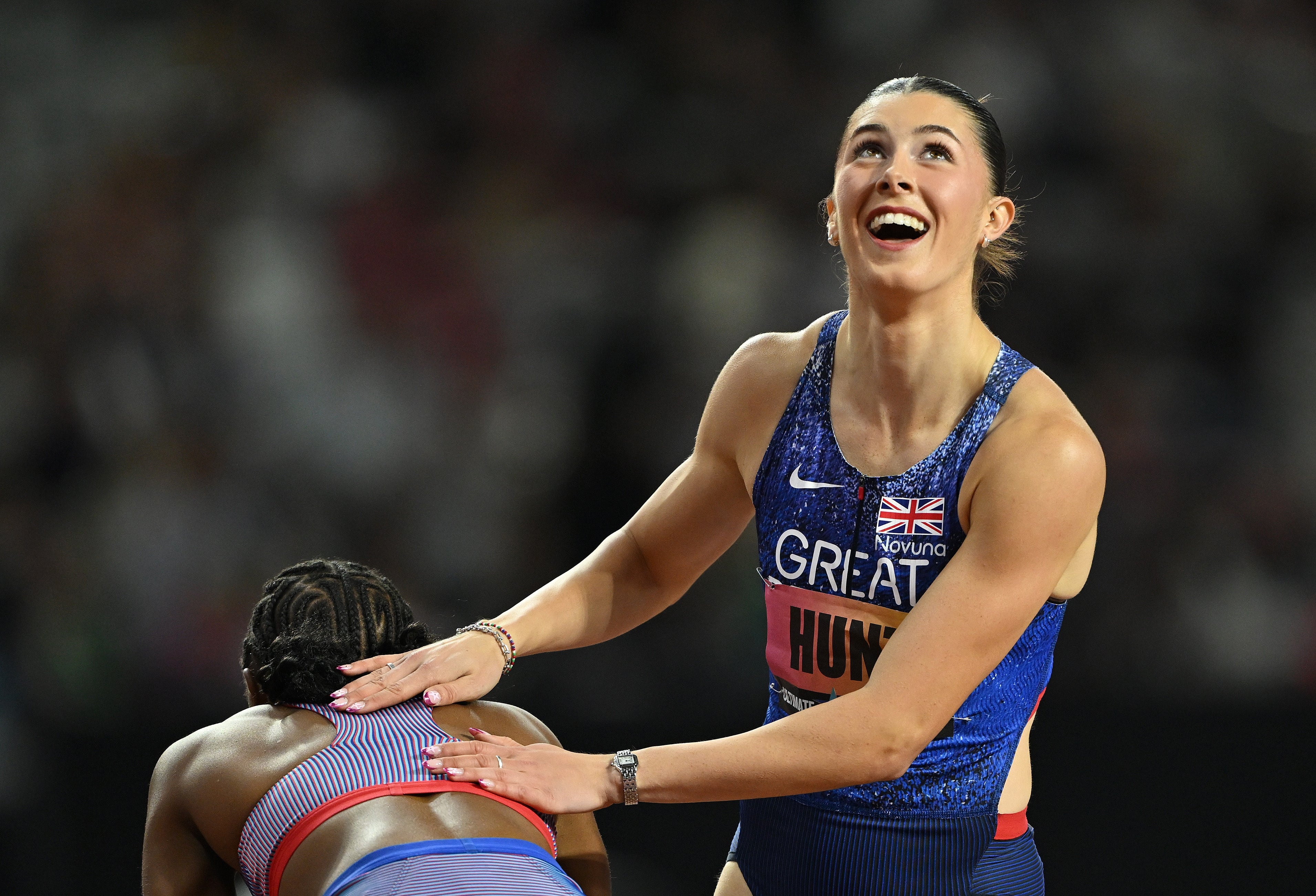 Idrettsutøver i atletisk utstyr, smilende og feirende på en bane, med en hånd på en medkonkurrents rygg