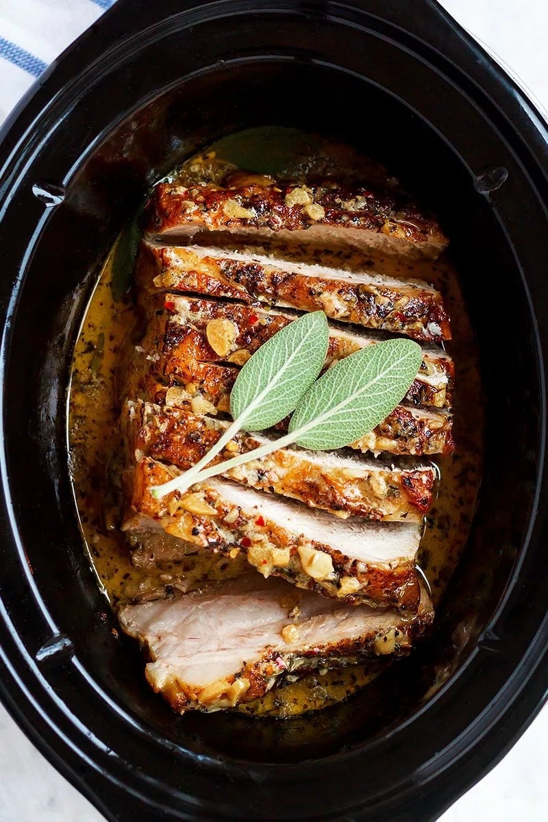 Sliced roast pork with herbs and garlic, garnished with sage leaves in a slow cooker
