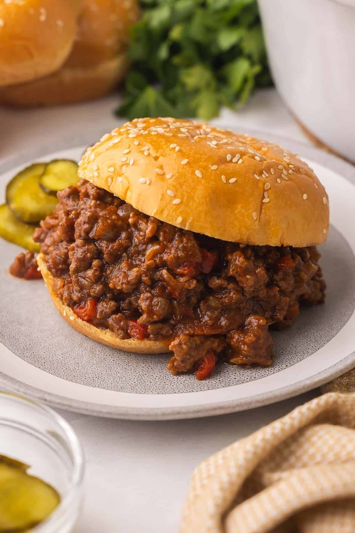Sloppy Joe sandwich on a sesame seed bun with a side of pickles on a plate