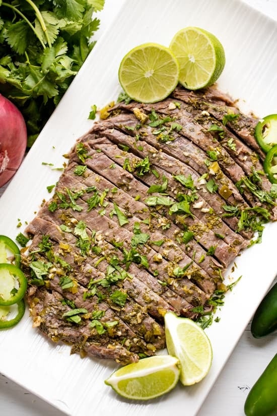 Sliced beef garnished with chopped herbs, lime wedges, and jalapeño slices on a white platter, next to cilantro and a red onion