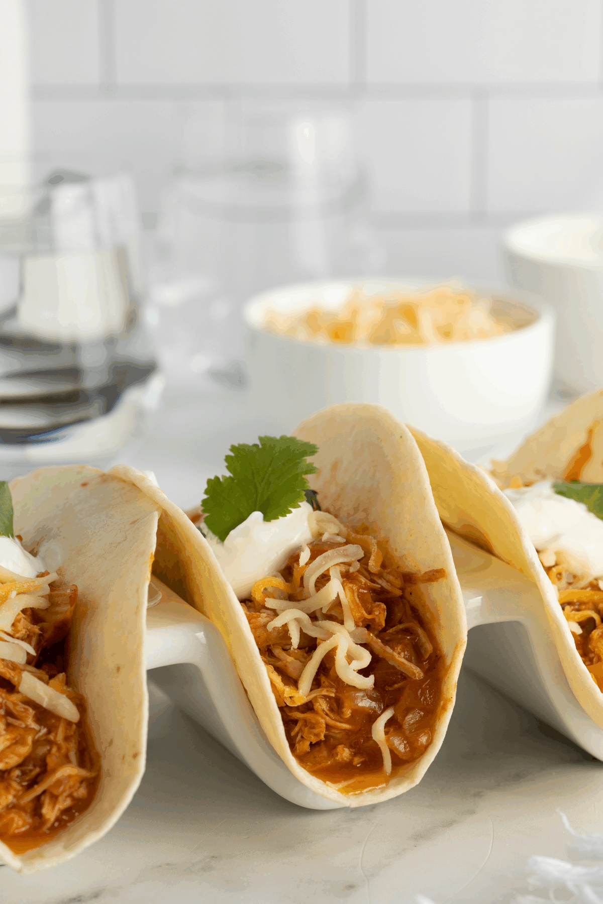 Three soft tacos filled with shredded chicken, cheese, and sour cream, garnished with cilantro, on a taco holder. Bowls in the background