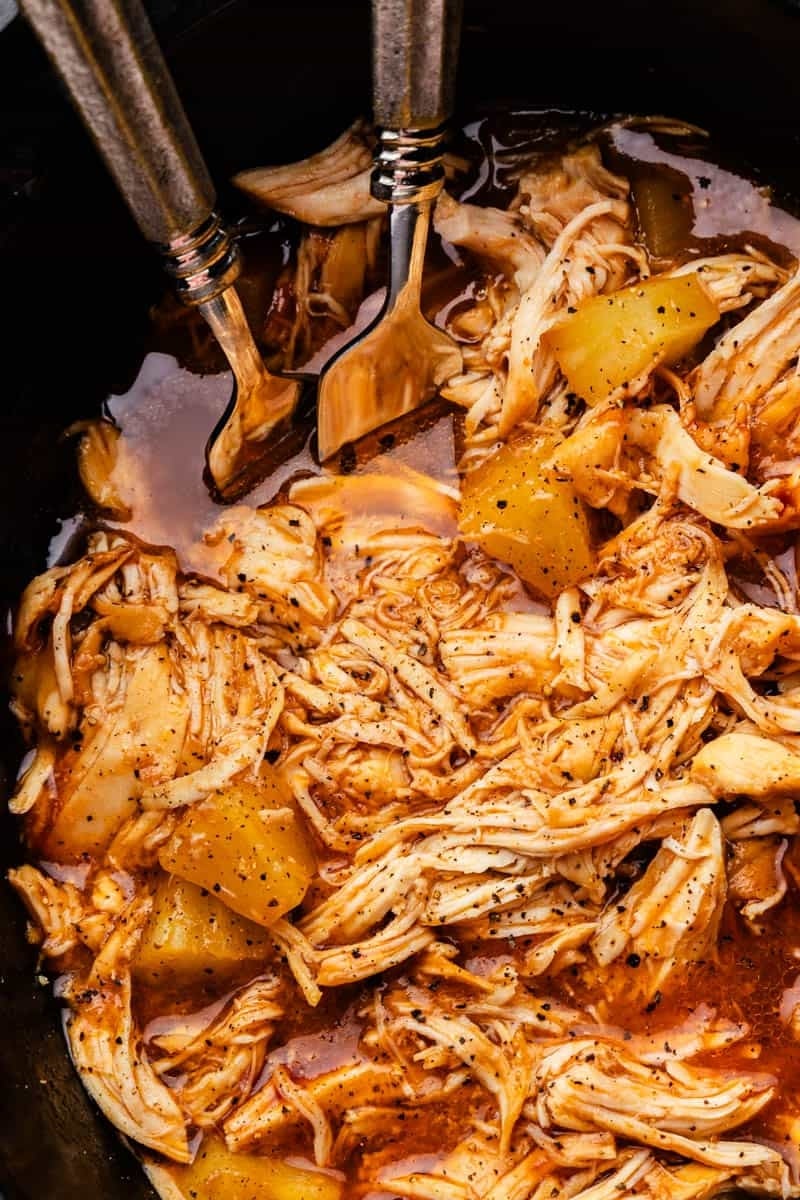 Shredded chicken with pineapple pieces and spices in a slow cooker, with two forks for serving