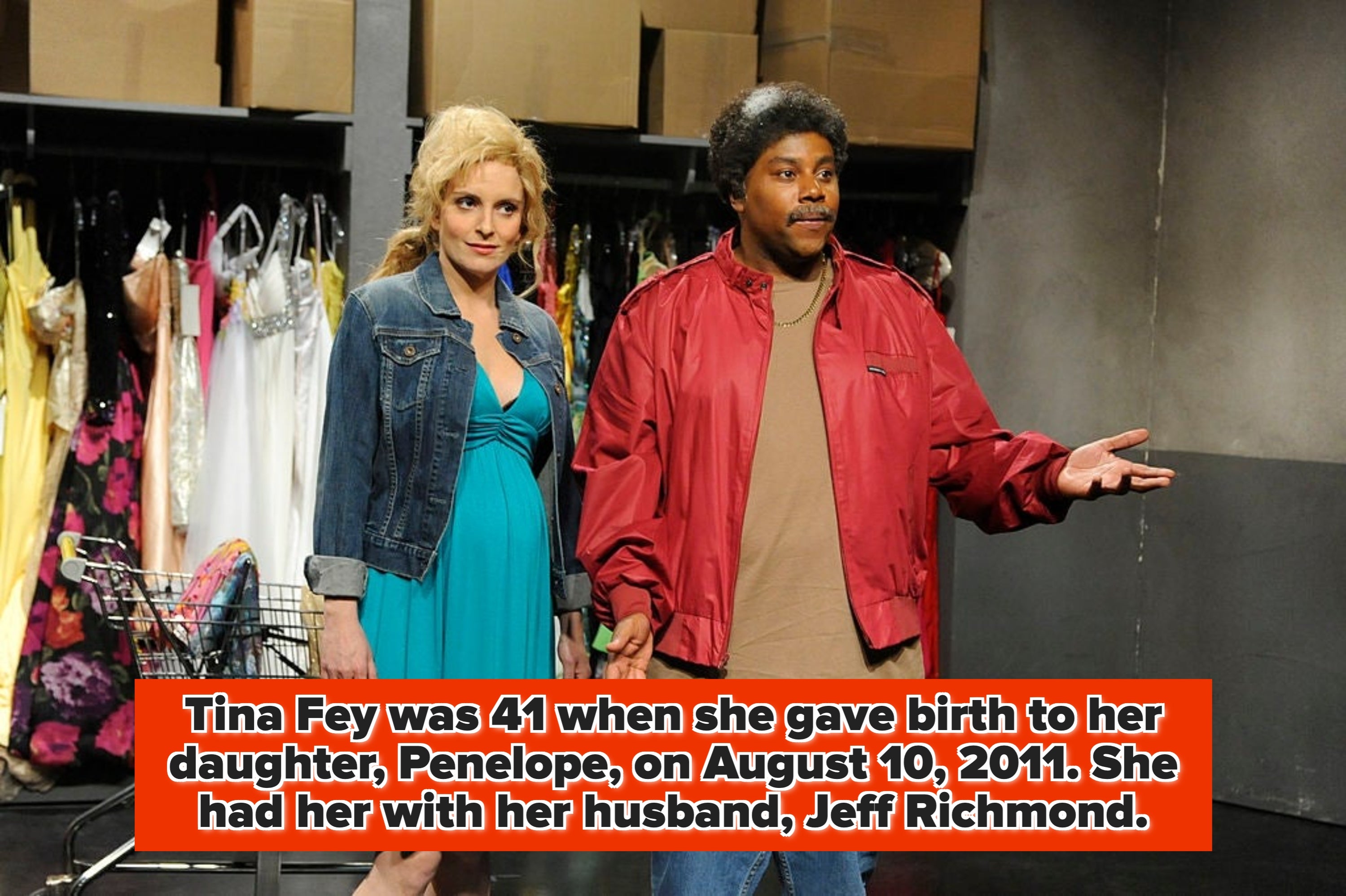 Two people on a set stand in front of a clothing rack. The woman wears a denim jacket and dress, and the man wears a red jacket