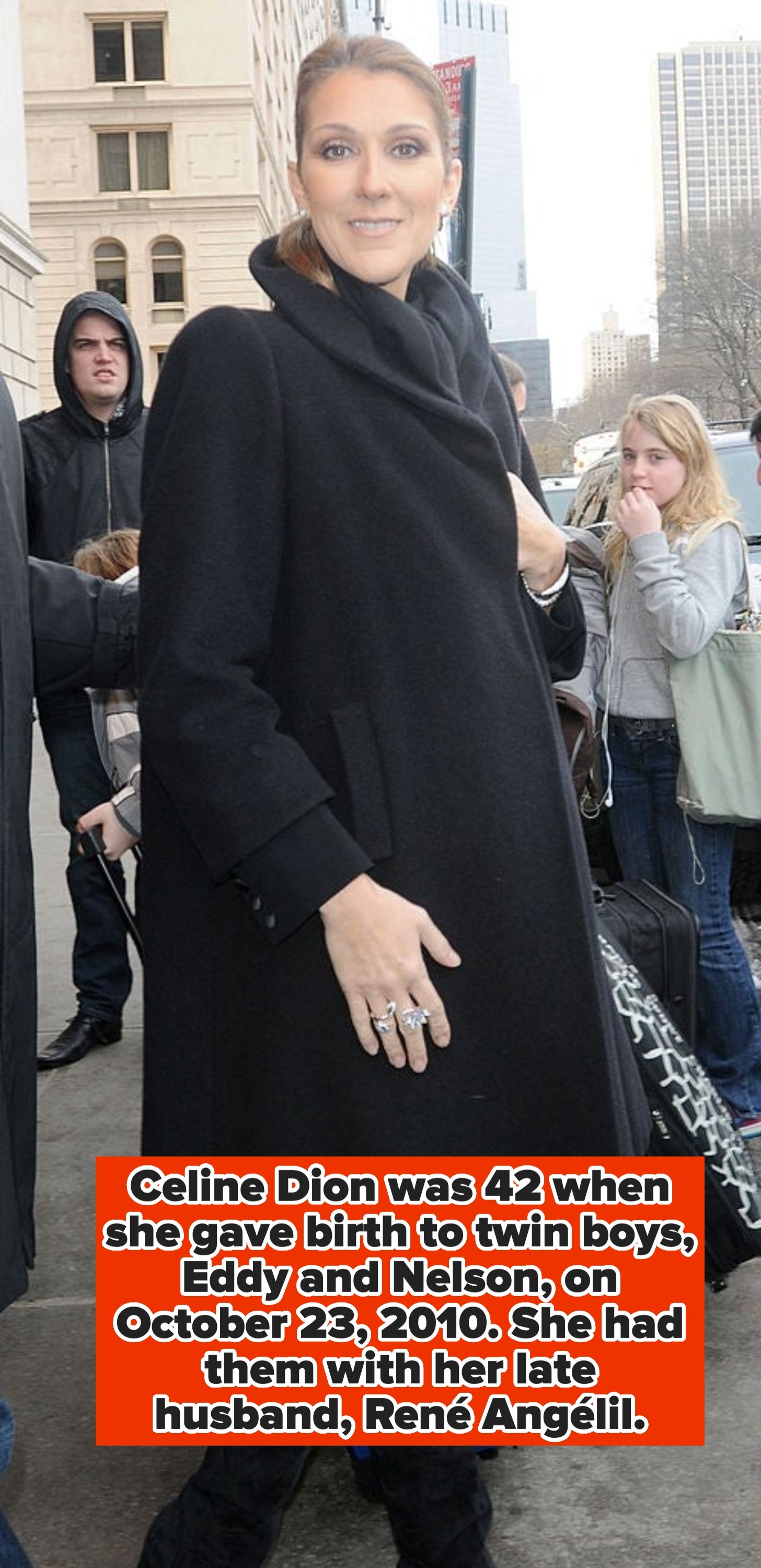 A celebrity in a chic, oversized coat with stylish rings and boots, surrounded by people in an urban setting
