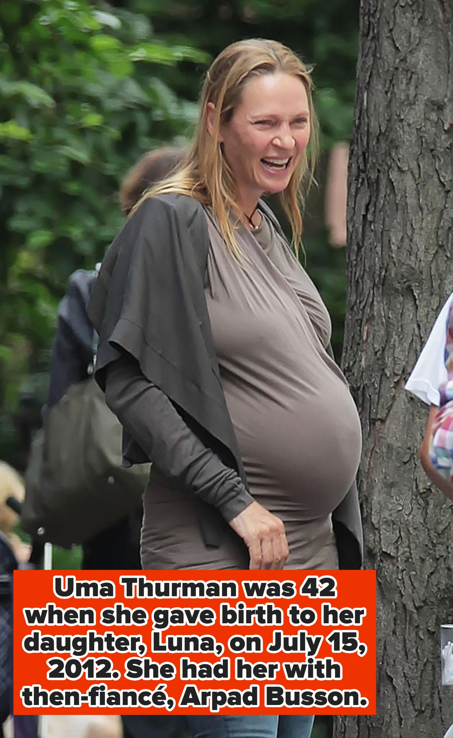 A smiling pregnant woman wears a casual top with long sleeves and jeans, standing outdoors near a tree