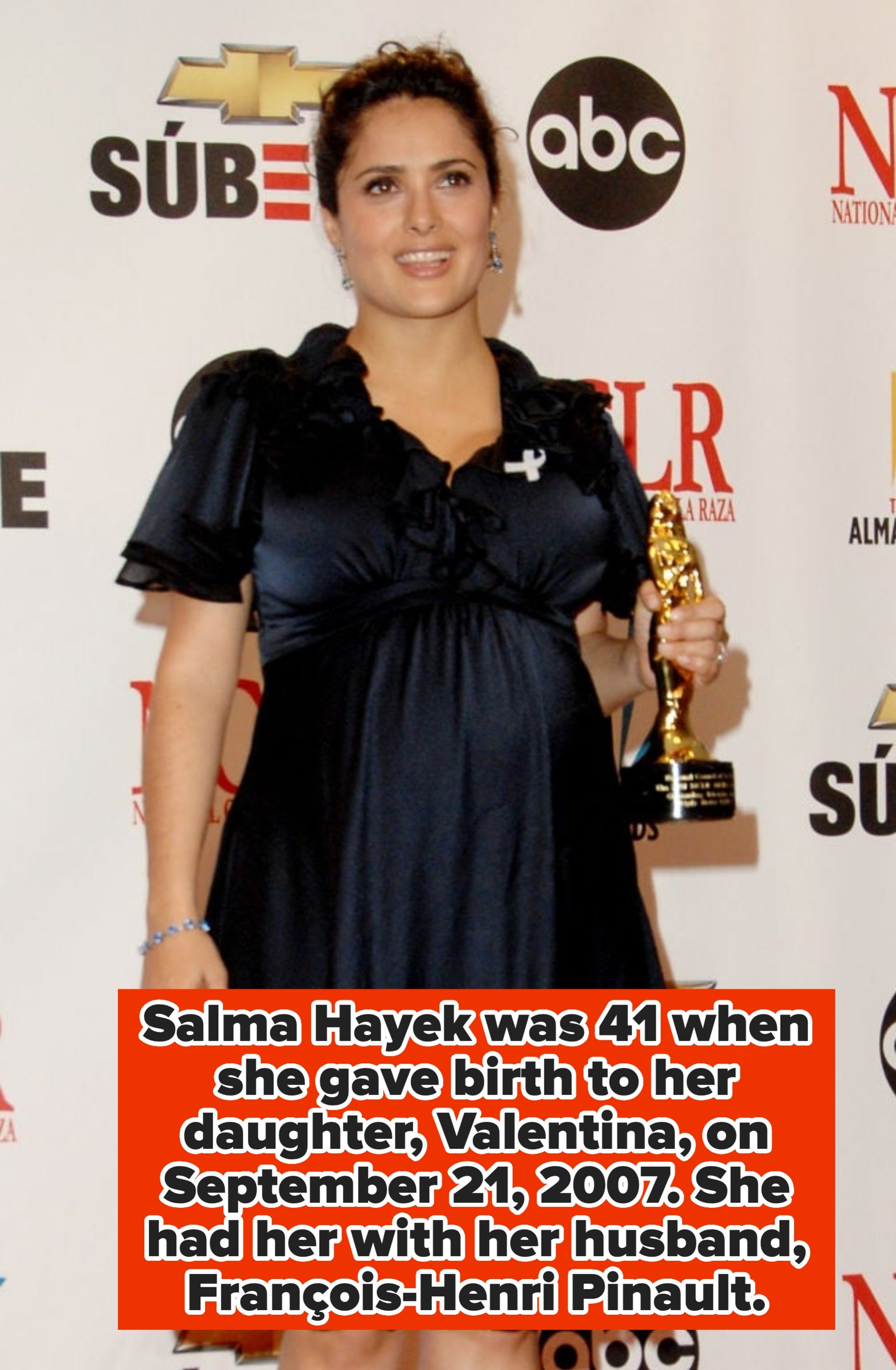Person in elegant dress holding an award on the red carpet, standing in front of a backdrop with logos