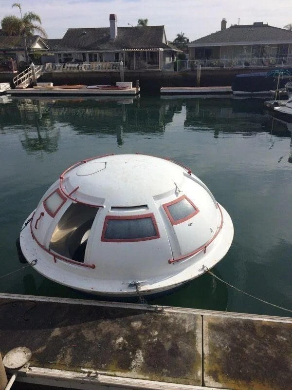A round, domed structure resembling a UFO is floating on water, tethered to a dock in a residential marina