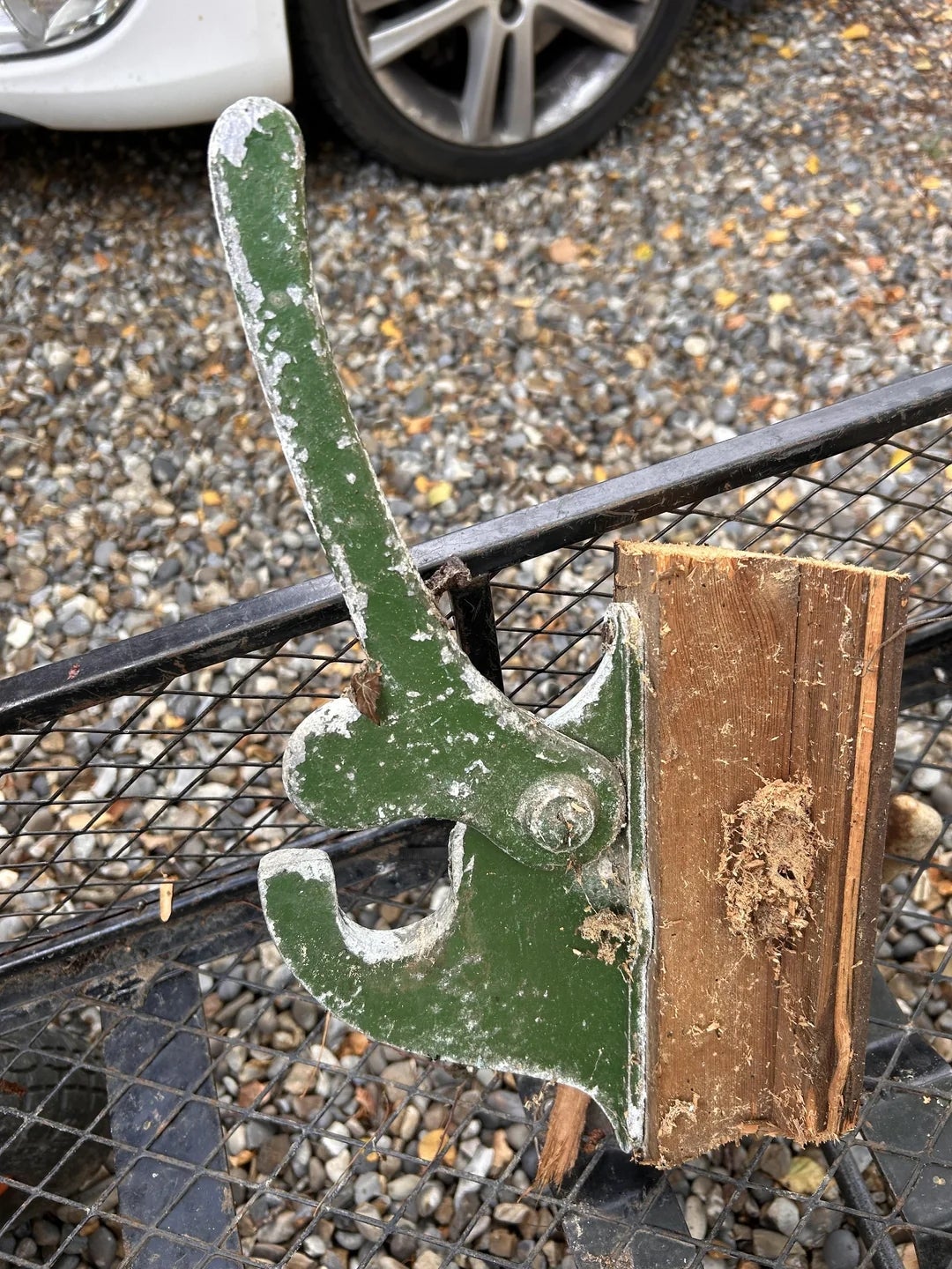 Old, weathered metal latch on wood, attached to a gridded metal frame, with a gravel surface and a car tire in the background
