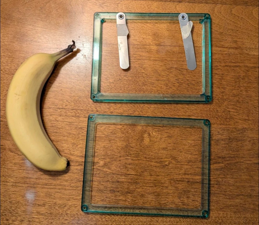 A banana next to a disassembled transparent rectangle with two metal hinges on a wooden surface