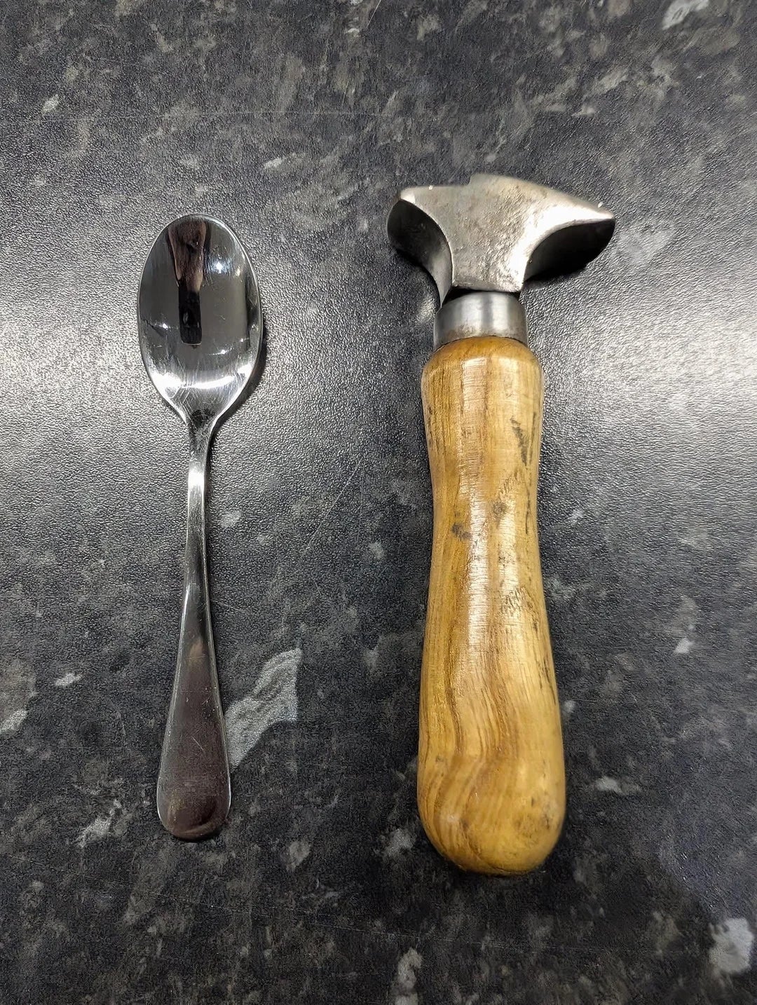 A small metal spoon next to a wooden-handled metal tool on a dark, textured surface