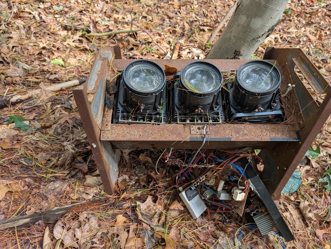 Three outdoor spotlights on a rusted metal frame, surrounded by fallen leaves and branches in a wooded area. Exposed wiring is visible below