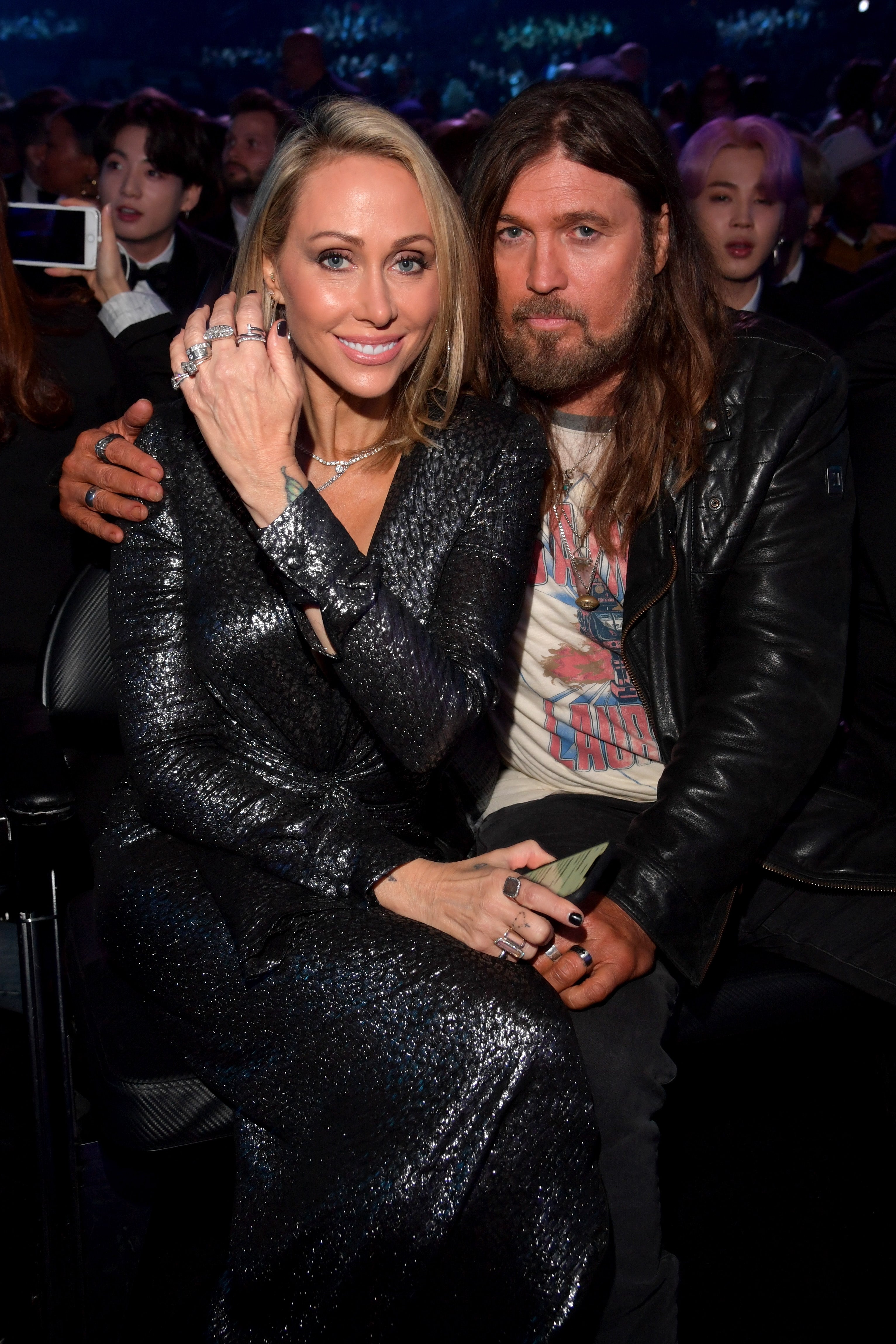 Tish Cyrus and Billy Ray Cyrus sit together at an event. Tish wears a shimmering dress, and Billy Ray wears a leather jacket and a printed T-shirt