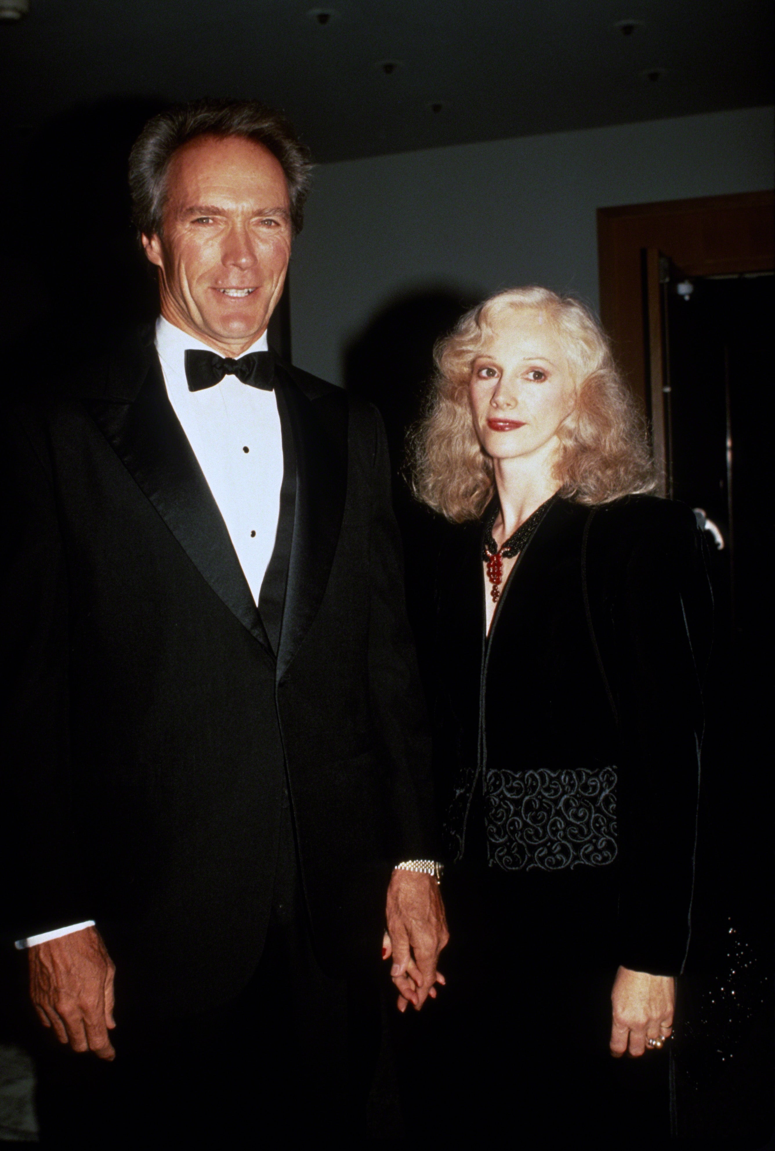 Clint Eastwood and Sondra Locke hold hands at an event. Eastwood wears a classic tuxedo and Locke wears an elegant, black dress
